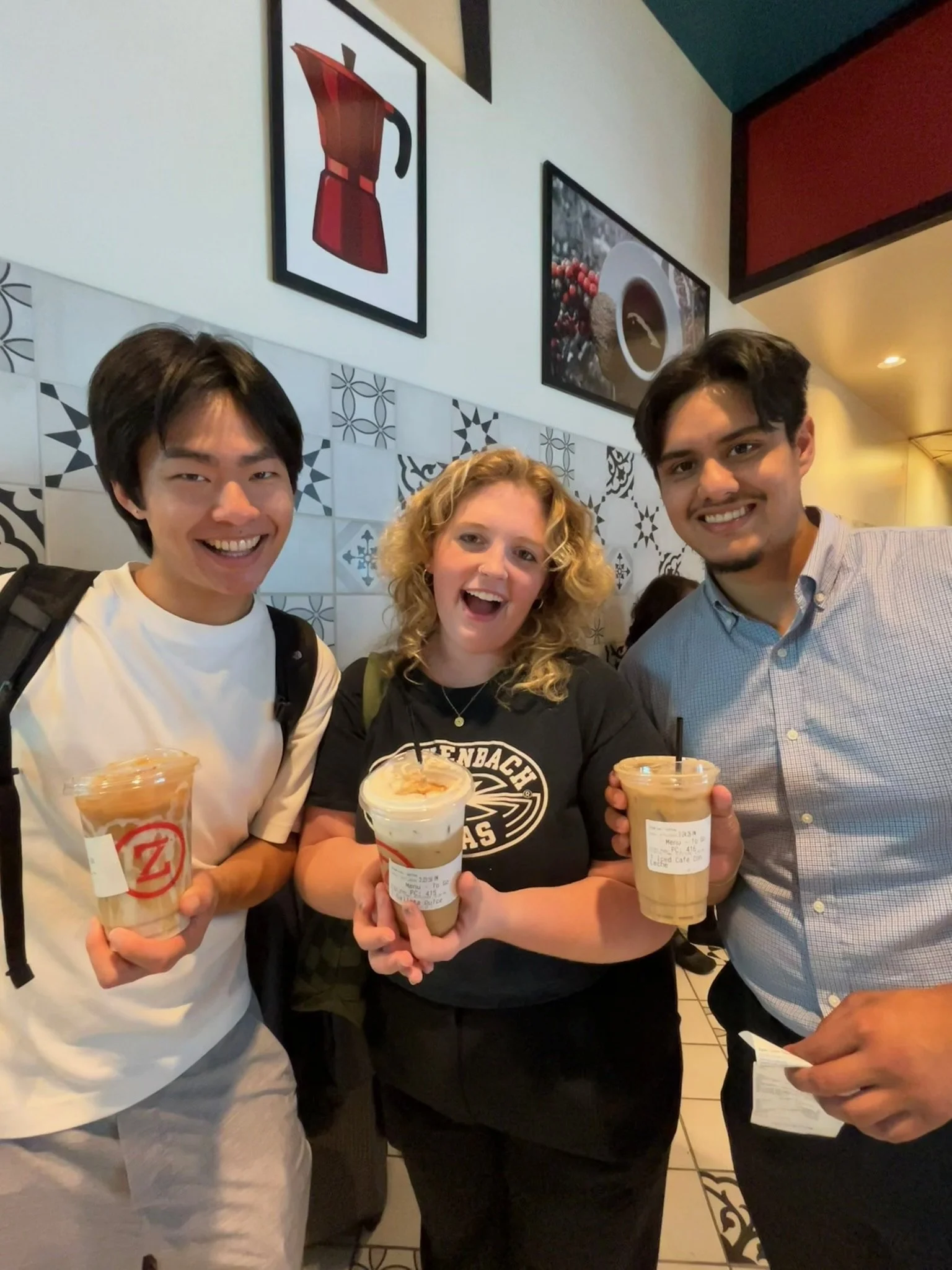 Three people smiling and holding drinks inside a cafe, with wall art depicting a coffee pot and a coffee cup behind them.