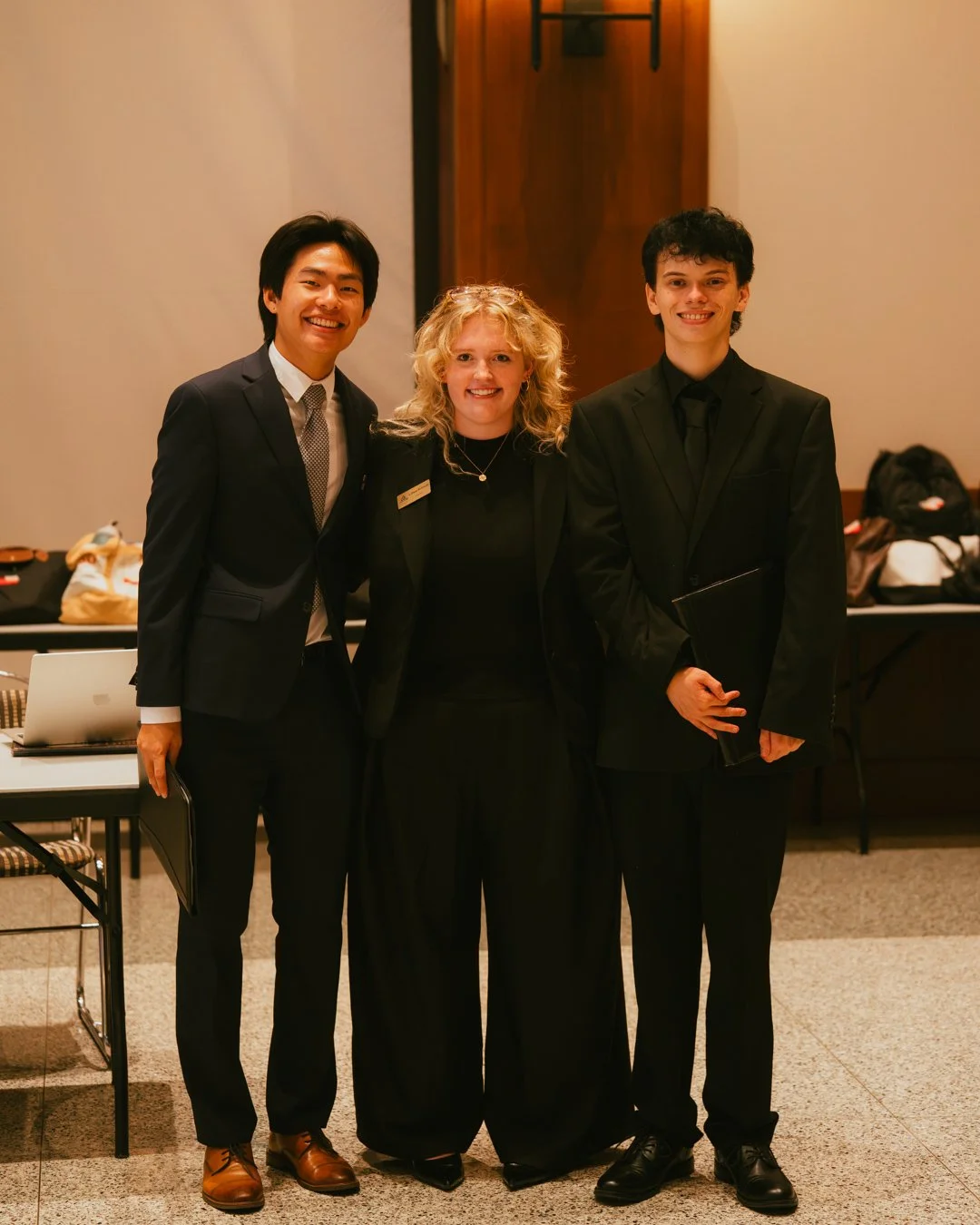 Three people in formal attire posing together indoors, with a table and bags in the background.