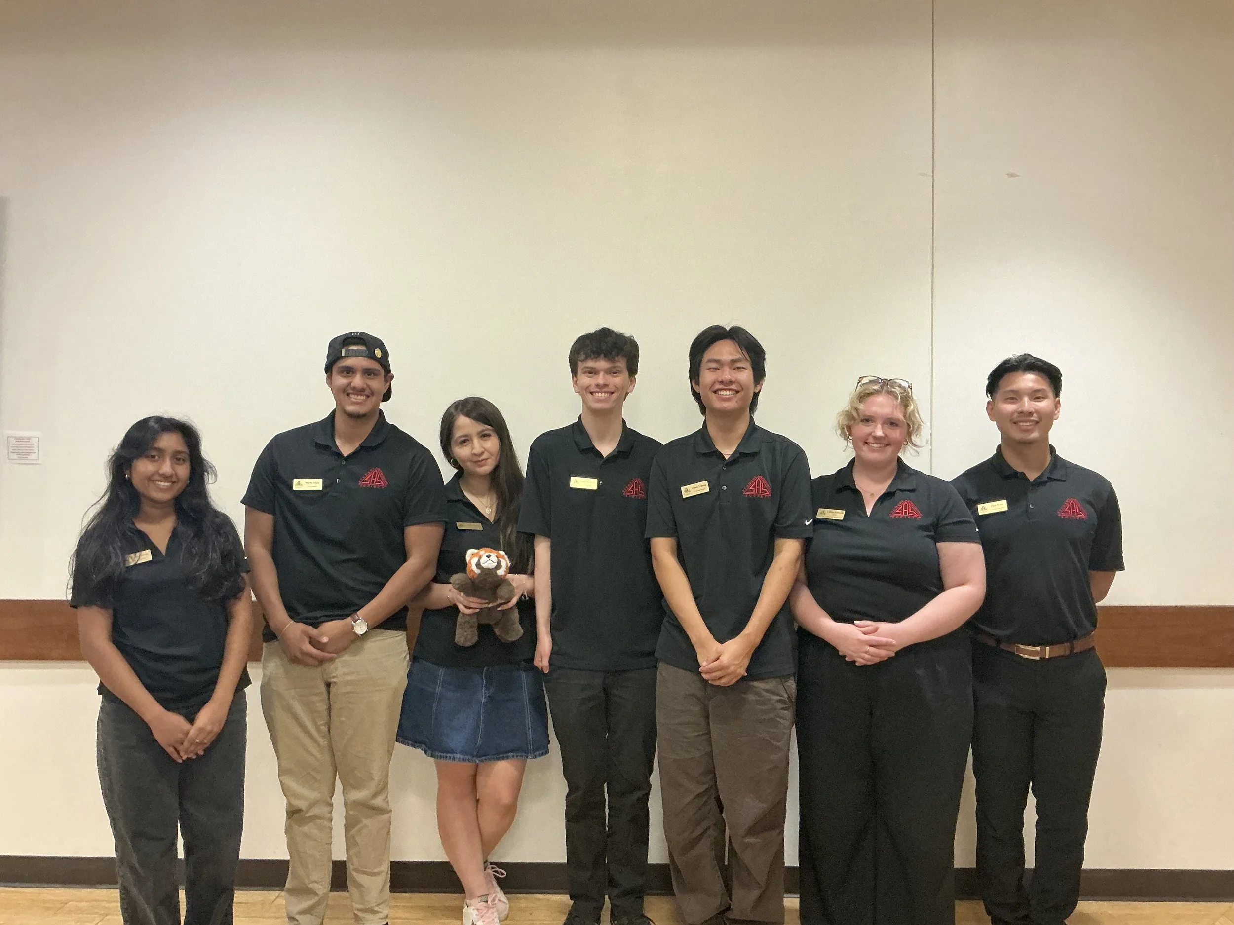 Group of seven young people standing together indoors, dressed in black polo shirts with red logos, smiling at the camera, with a plain beige wall in the background.