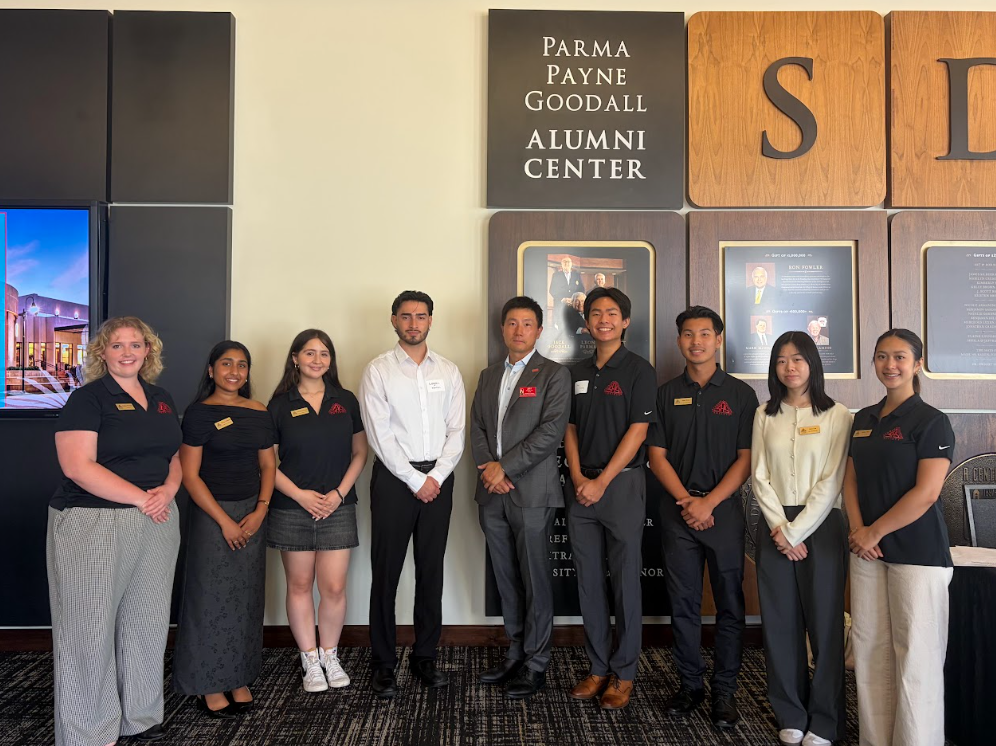 Group of nine people standing in front of a wall with plaques at Parma Payne Goodall Alumni Center.