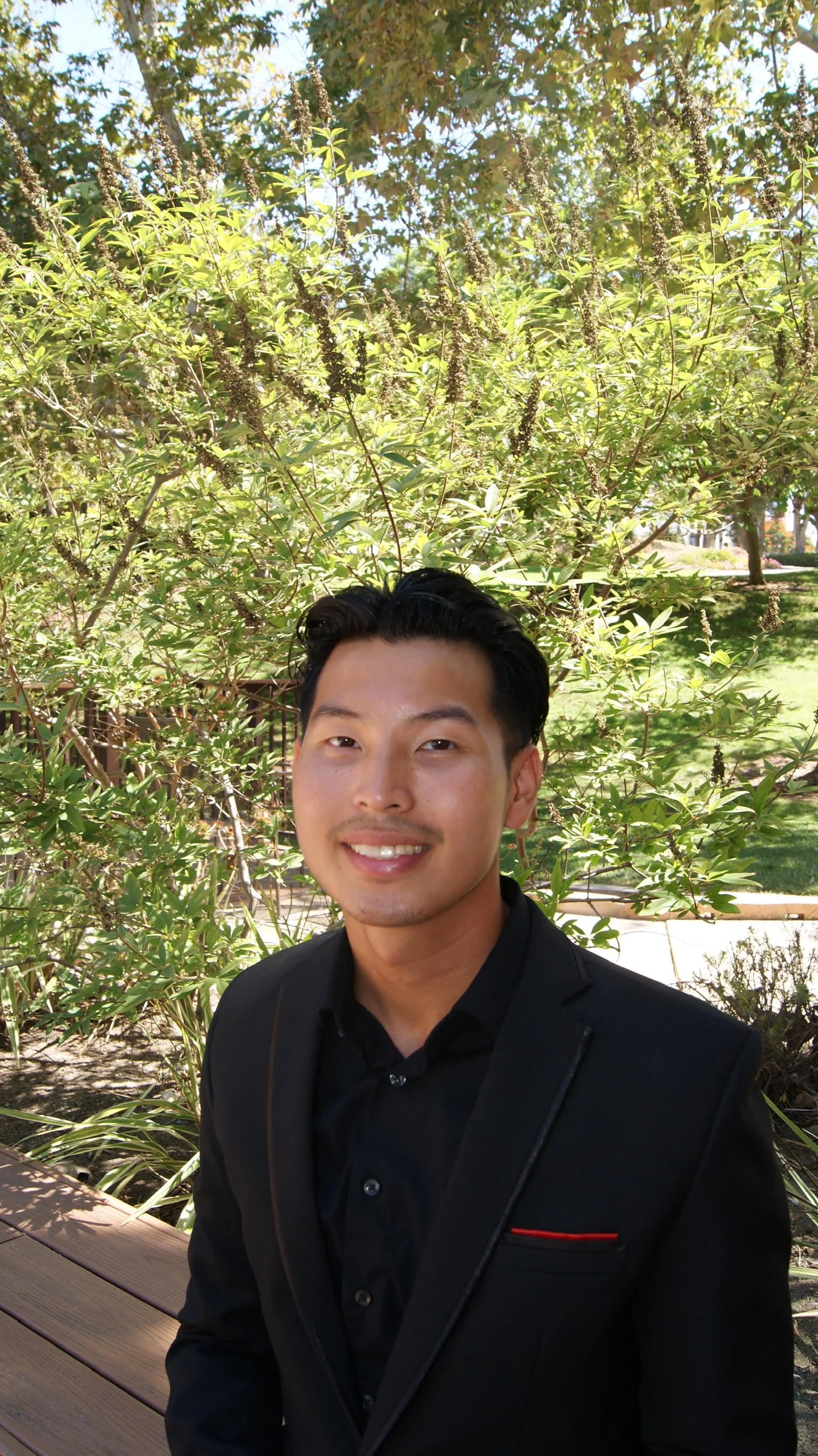 A young man with dark hair and a smile, wearing a black shirt and blazer, standing outdoors in front of green foliage and plants.