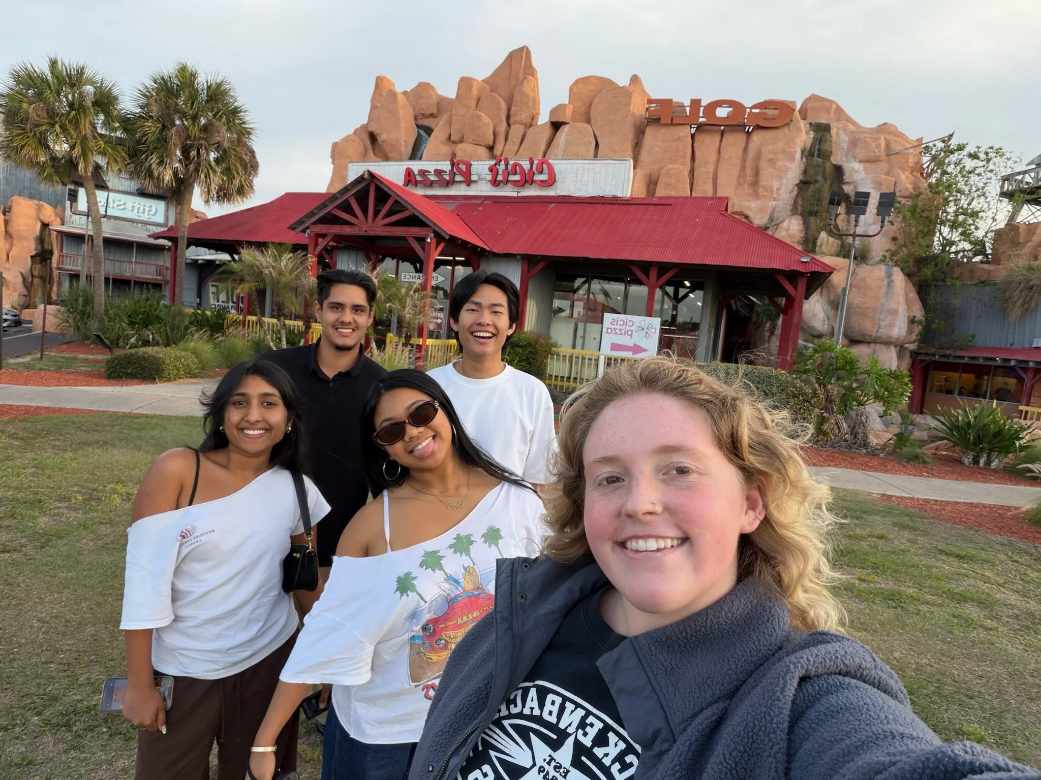 Group of five young adults taking a selfie in front of a rock formation with signage at a themed amusement park, including a sign for a roller coaster and a building with red roof.