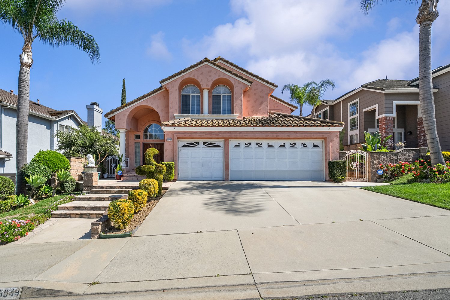 A two-story Chino Hills house with a tiled roof, white garage doors, and tropical landscaping, including palm trees and neatly trimmed bushes, under a partly cloudy sky.