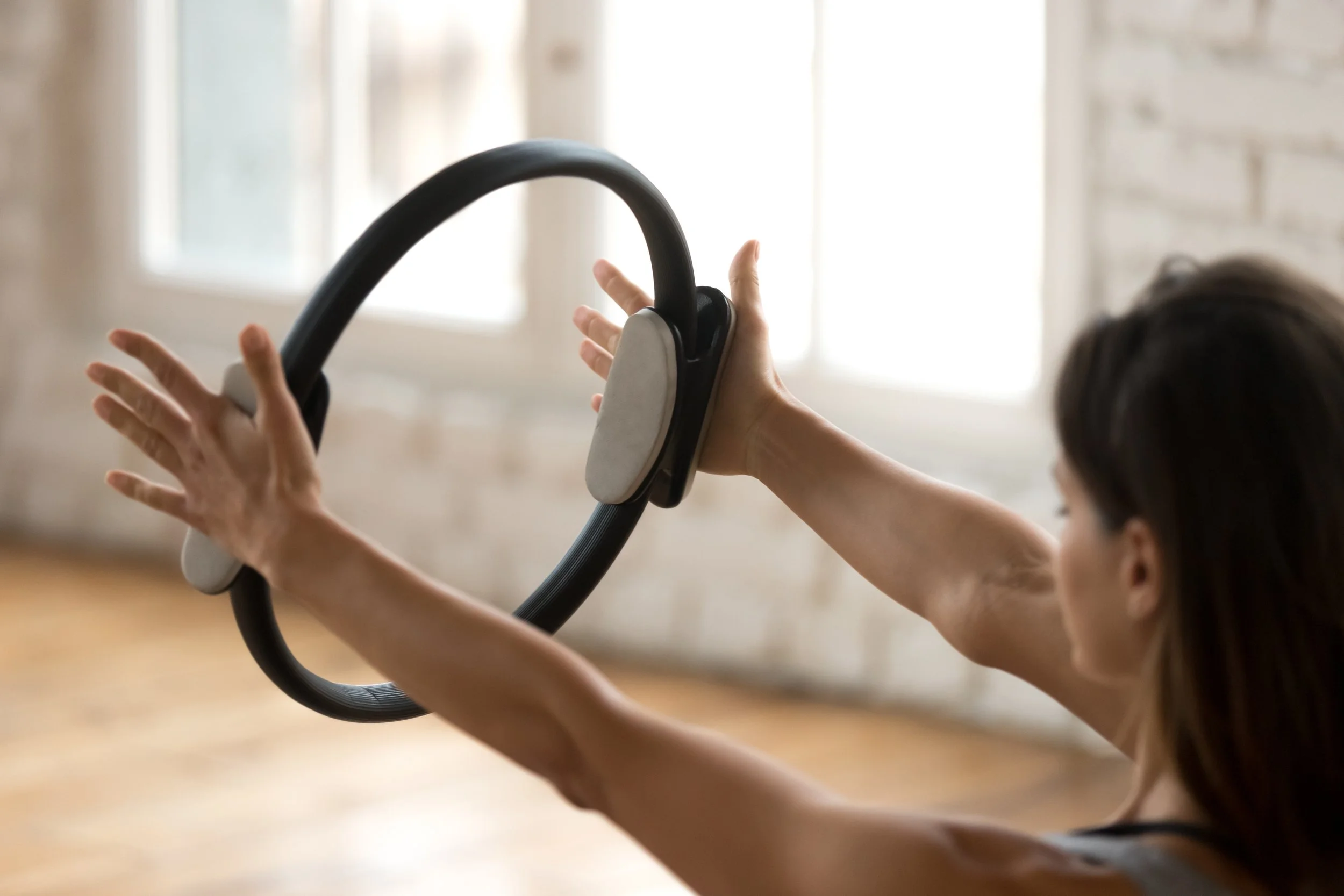 A person holding a Pilates magic circle, with their hands holding the outsides of the magic circle, in a room with windows and wooden floors.