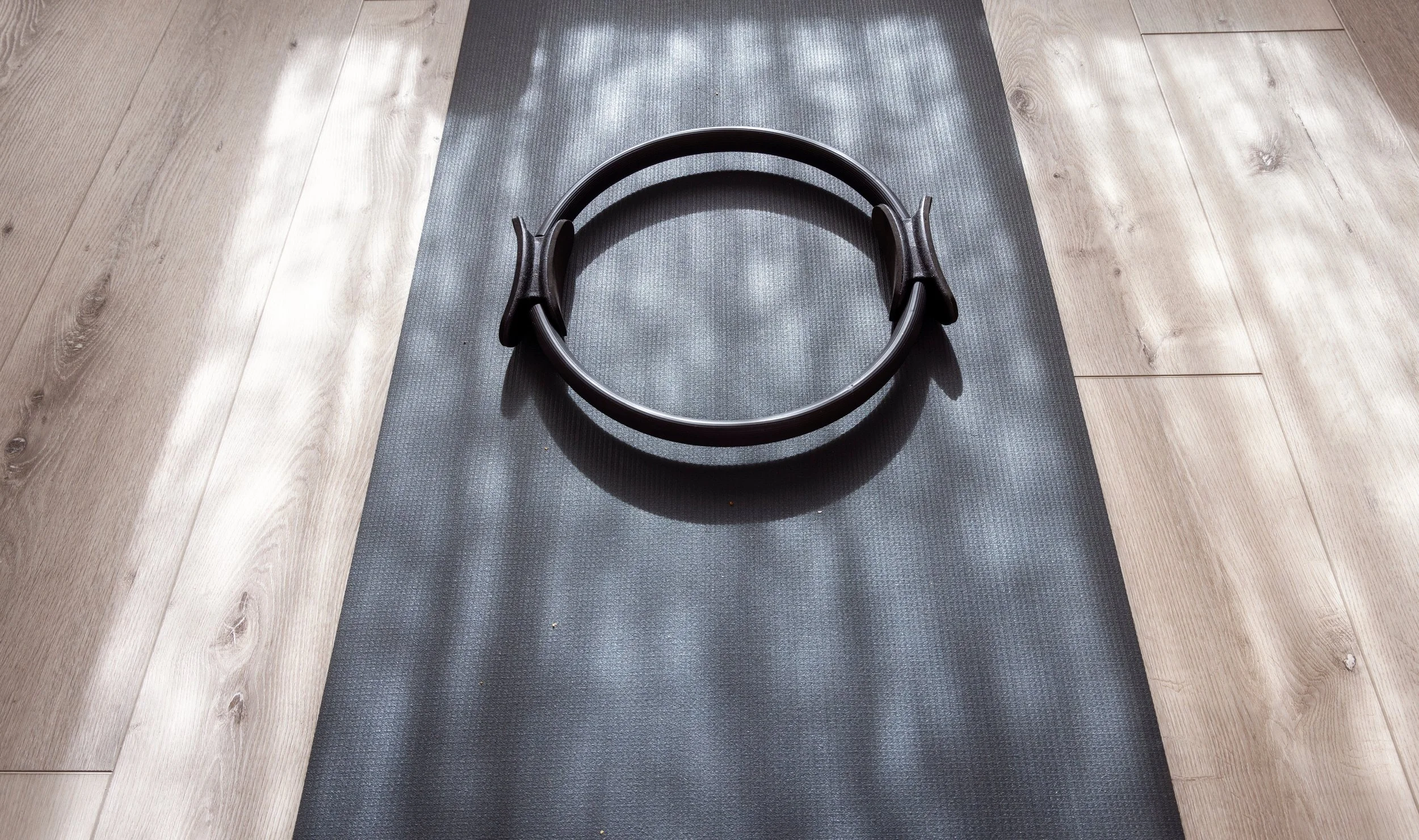 A black Pilates Magic Circle placed on a dark gray workout mat, with a light-colored wooden floor surrounding the mat.