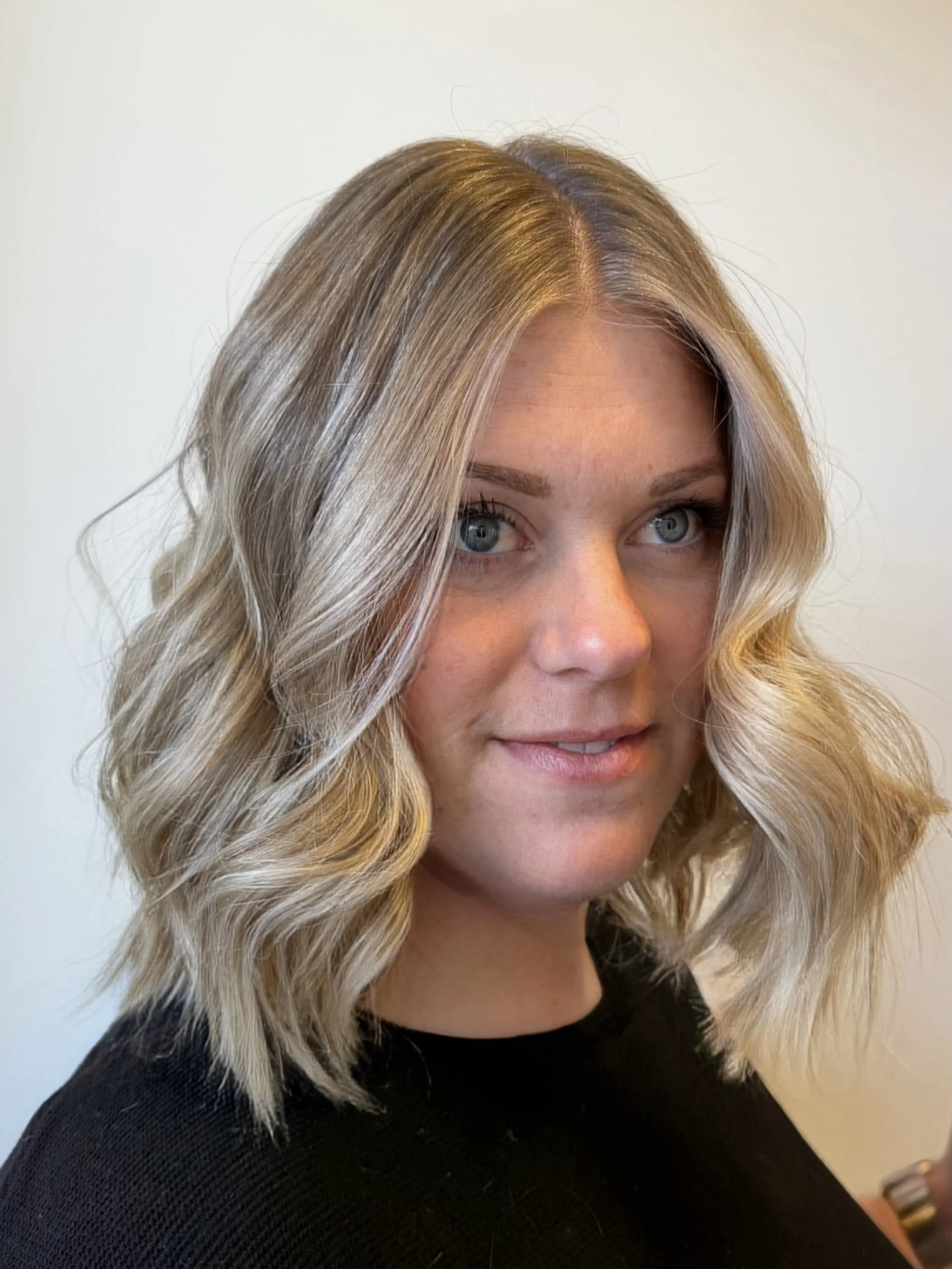 A woman with wavy blonde hair and blue eyes wearing a black top, smiling slightly against a plain white wall background.