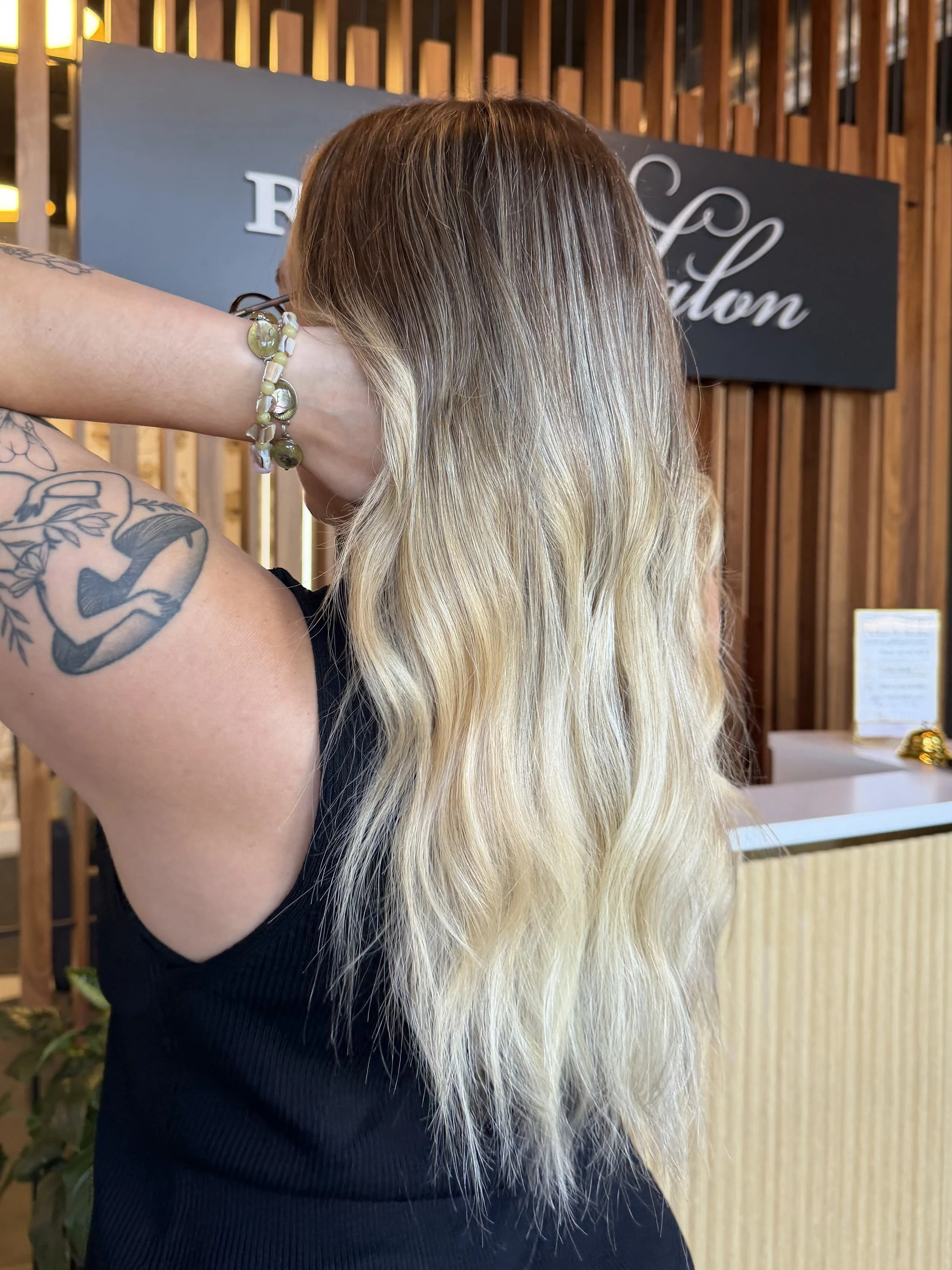 A woman with long, wavy blonde hair crossing her arm behind her neck. She has a sleeve tattoo on her upper arm and a beaded bracelet. She is wearing a black sleeveless top and is standing in front of a wooden decor with a sign that reads 'R + Carlton'.