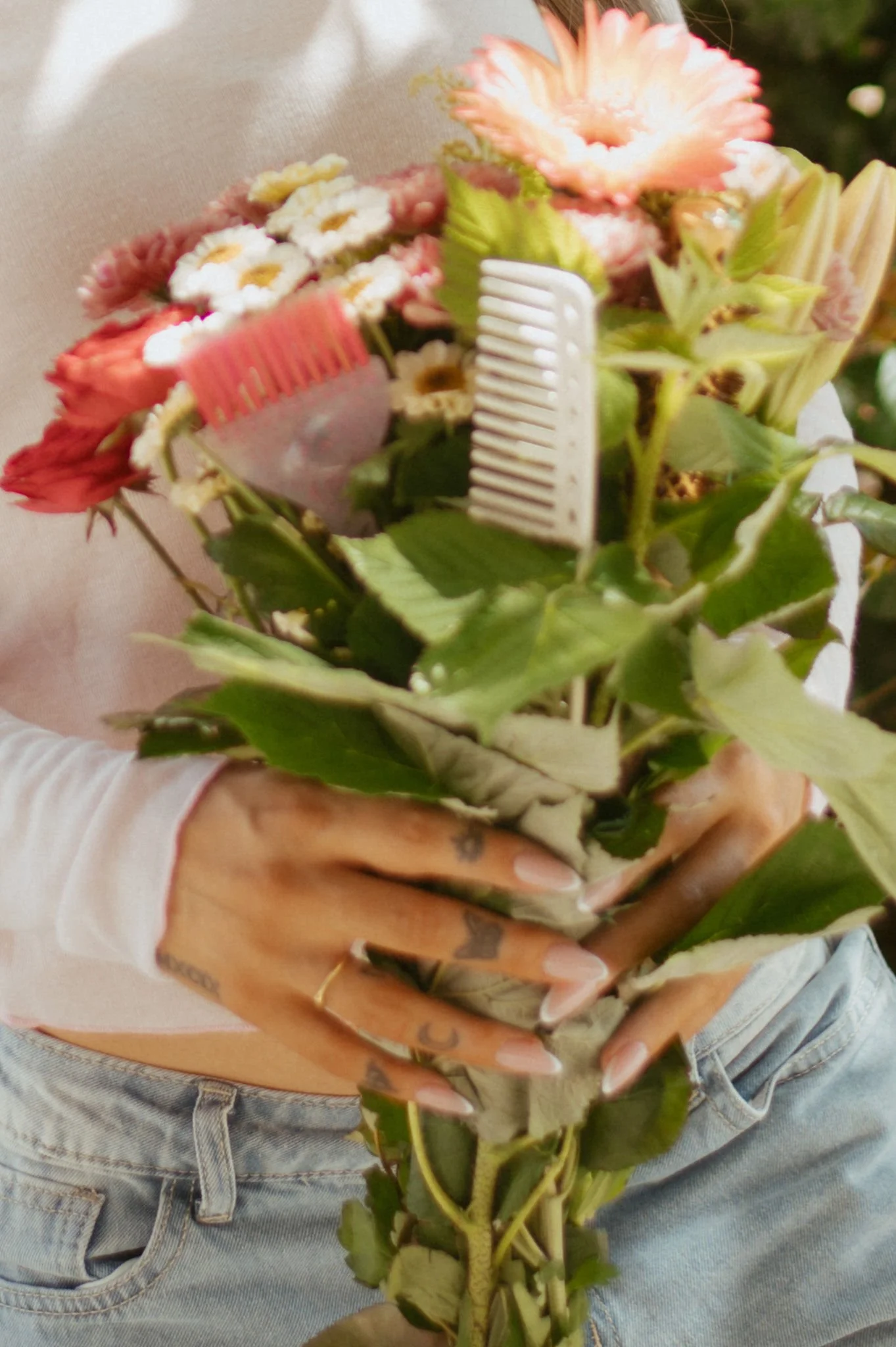 Person holding a bouquet of colorful flowers, including pink and white daisies, with a beige background and wearing a white long-sleeve shirt and light blue jeans.