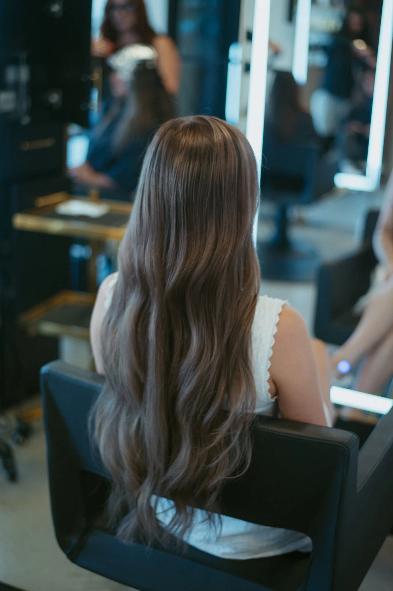 A woman with long wavy brown hair sitting on a black chair, facing a mirror in a salon, with other people visible in the background.