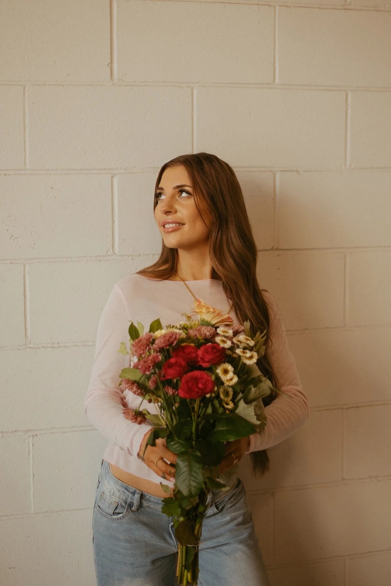 A young woman with long brown hair holding a bouquet of various colored flowers, standing against a white brick wall and wearing a light-colored shirt and jeans.