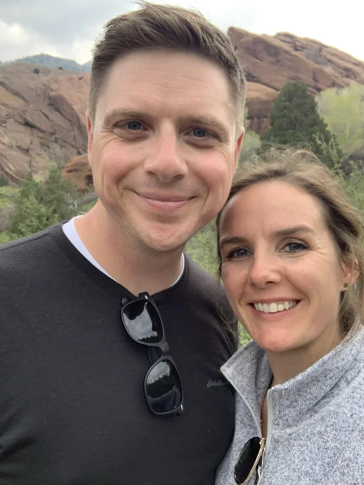 A smiling man and woman outdoors with rocky hills and greenery in the background.