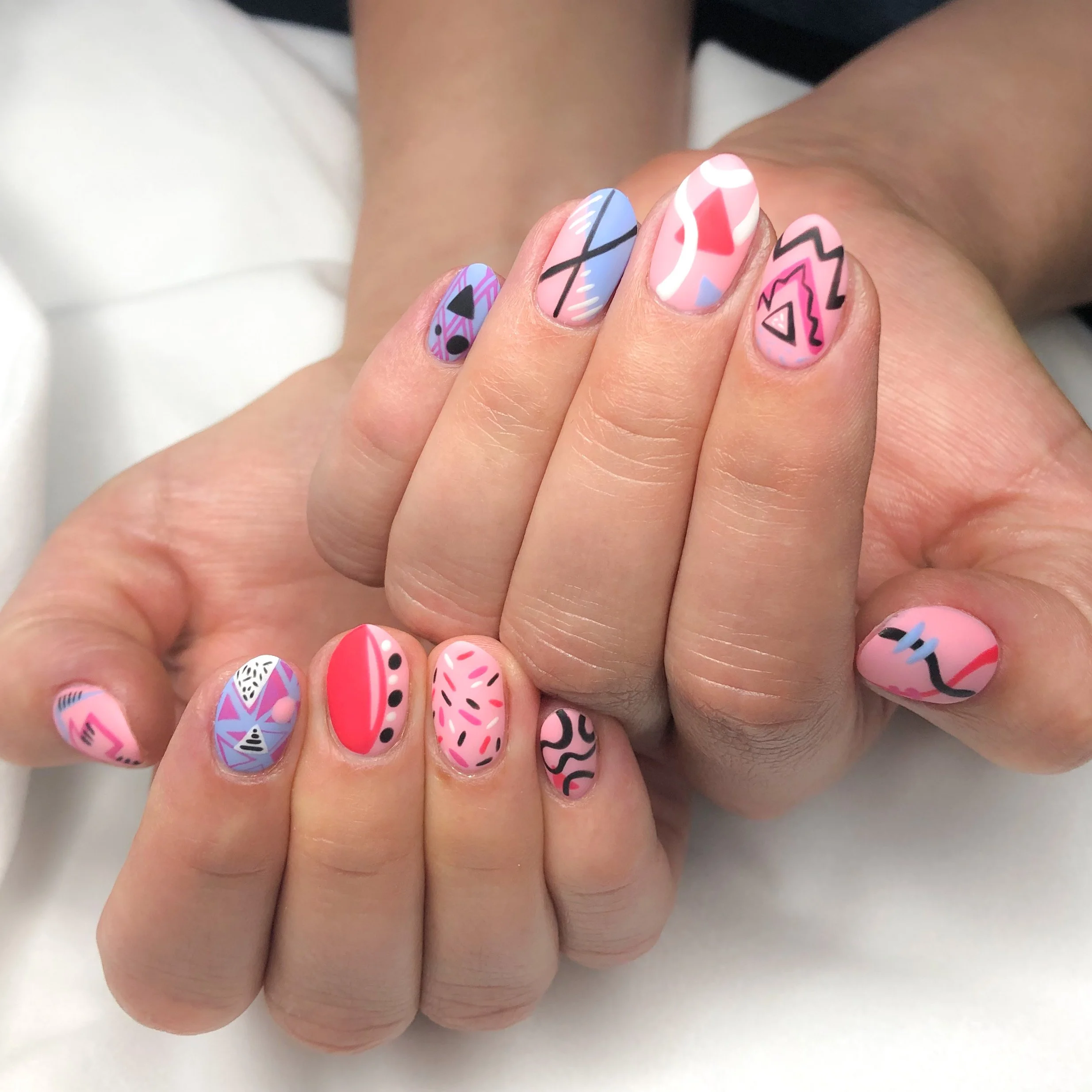 Close-up of fingers with colorful, abstract, geometric nail art designs in shades of pink, purple, blue, black, and white.