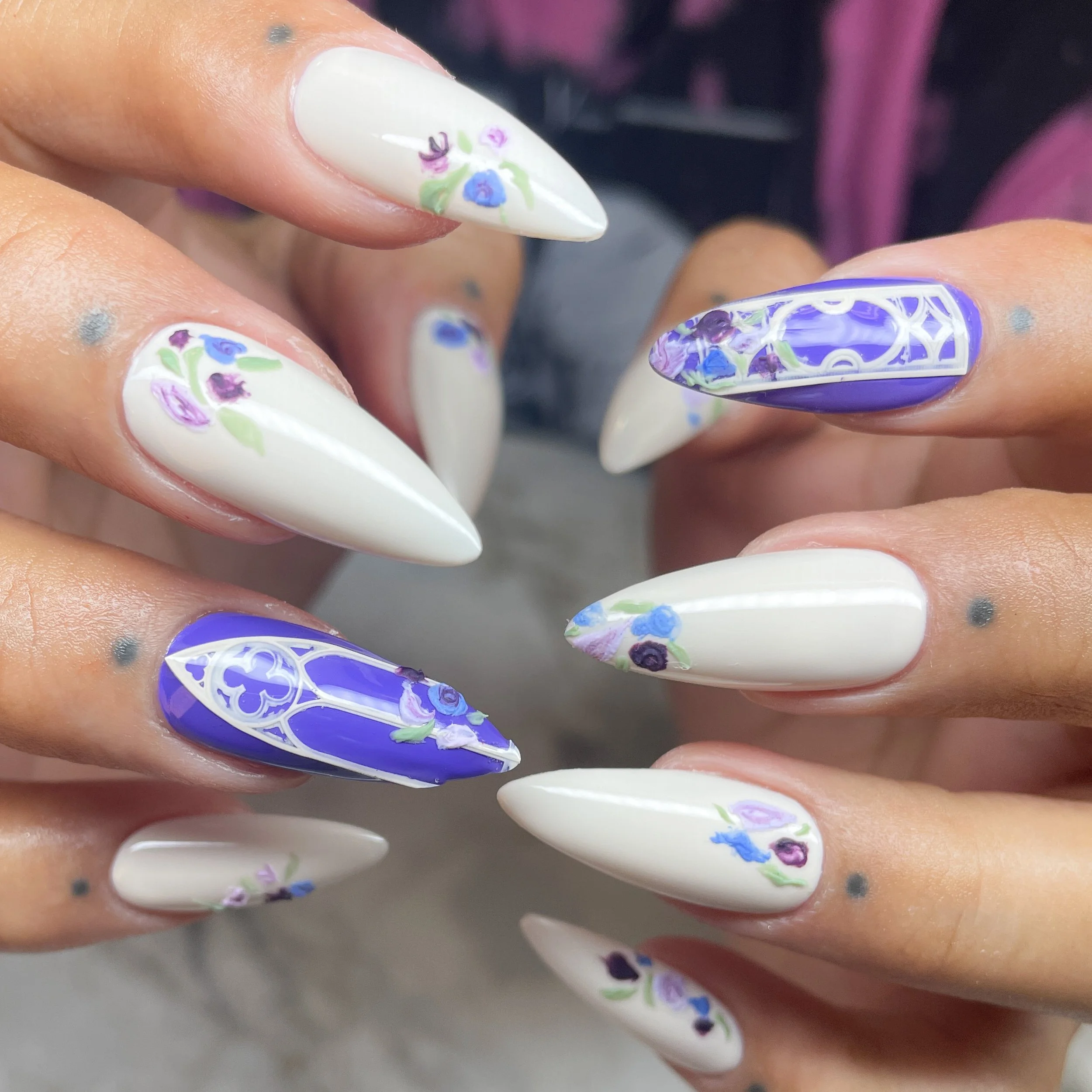 Close-up of manicured nails with floral and geometric nail art designs in white, purple, blue, and green colors.
