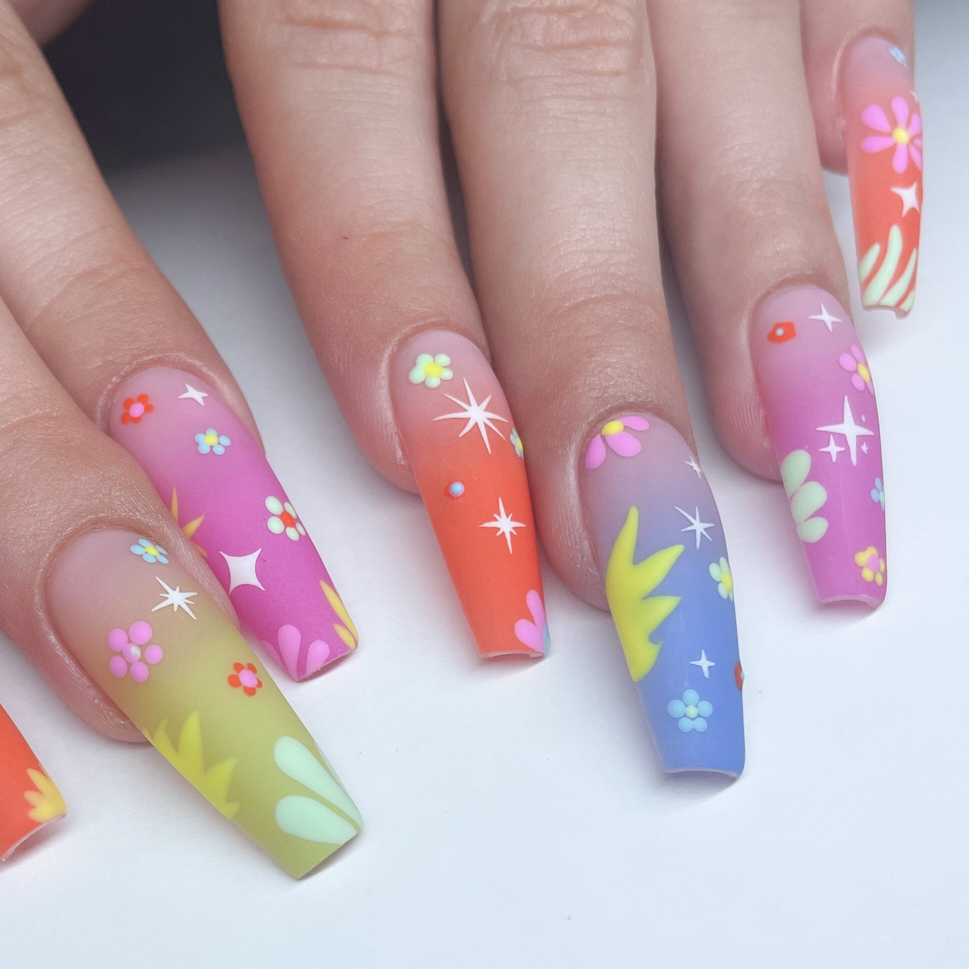 Close-up of decorated fingernails with pastel pink, orange, yellow, green, and blue gradient base colors, accented with small flower and star-shaped nail art designs.