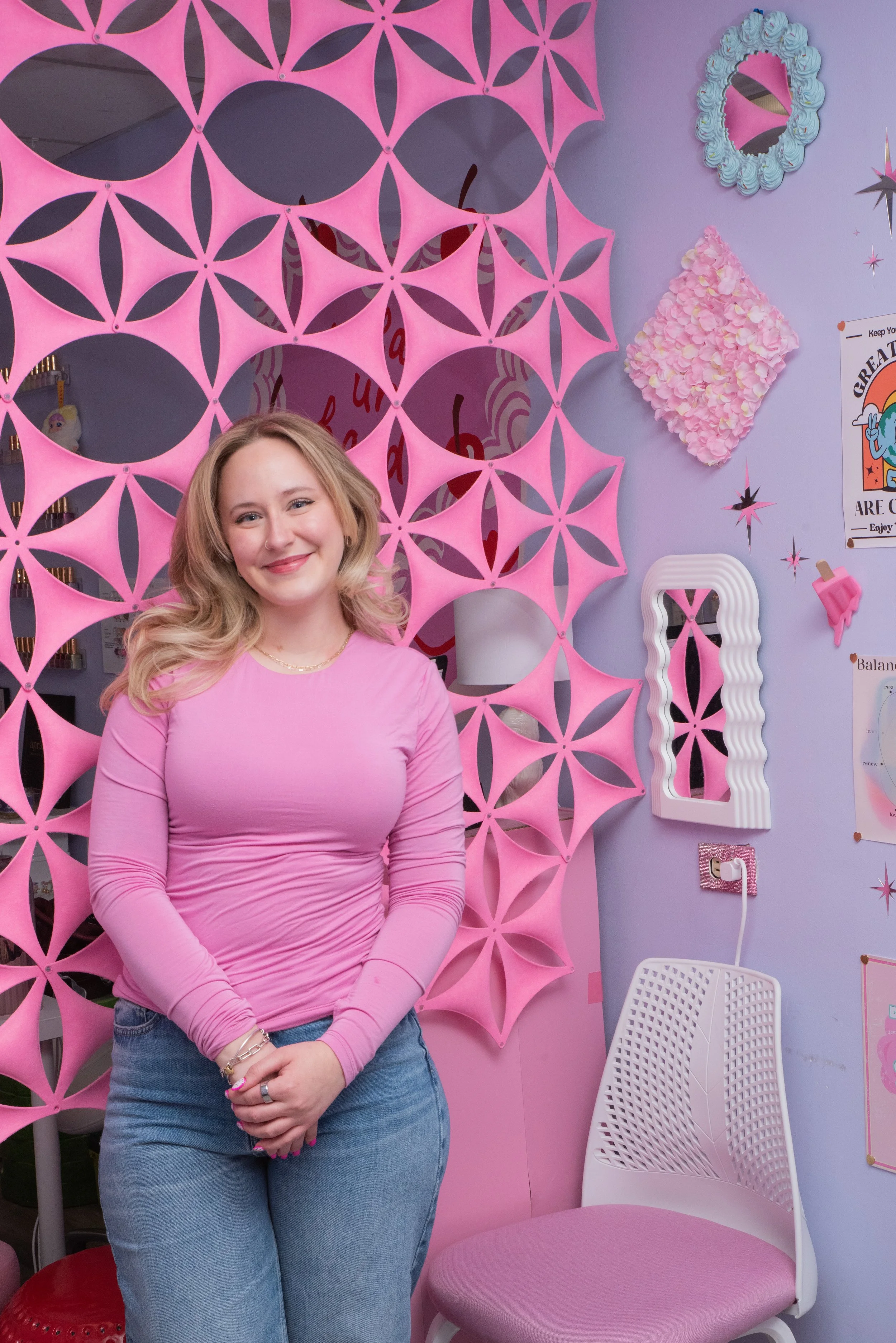 A young woman with blonde hair smiling, wearing a pink long-sleeve shirt and jeans, standing in front of a pink decorative wall with circular cutouts and star shapes, and various wall decorations including a mirror, a pink floral art piece, and posters.