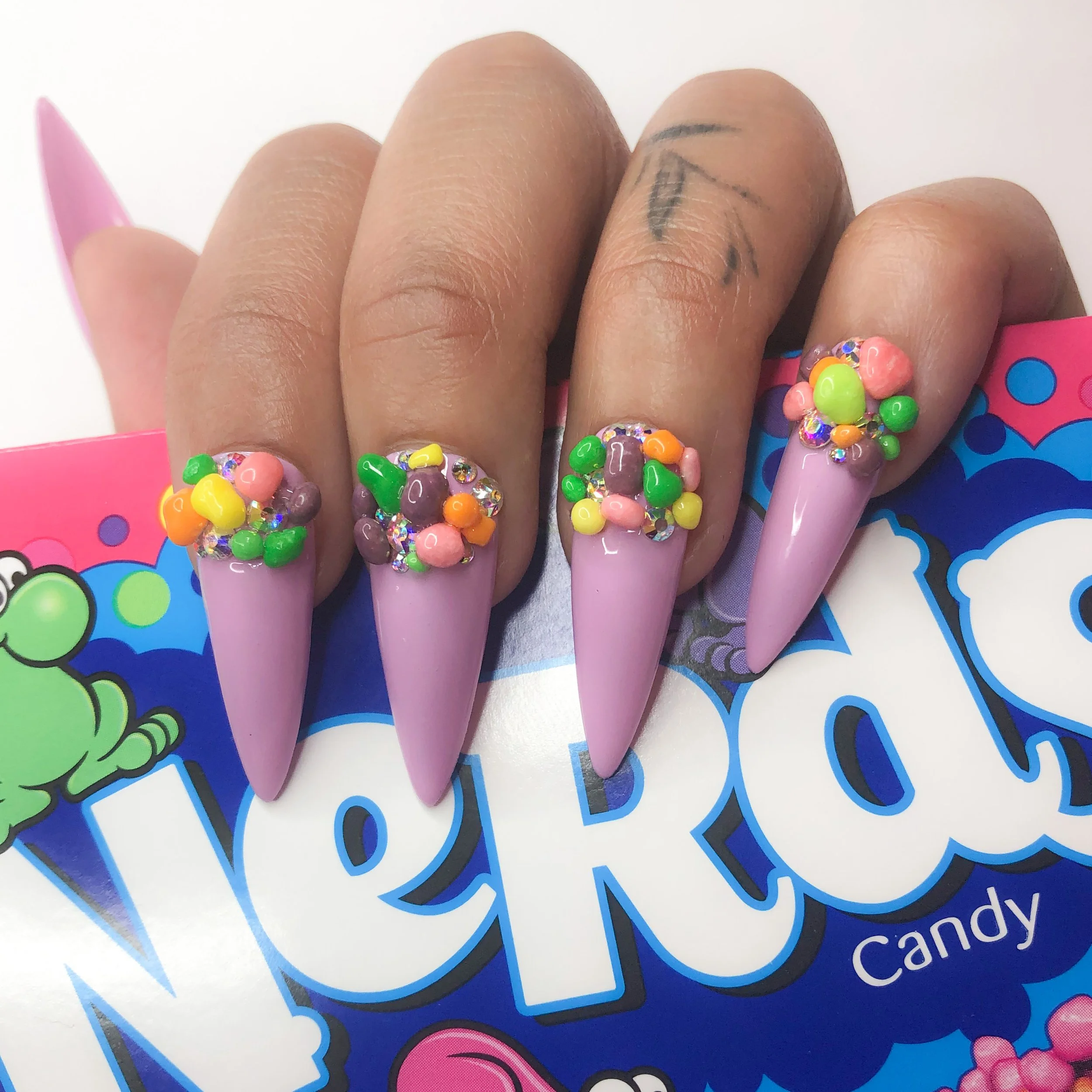Close-up of a person's hand with colorful 3D nail art decorated with small beads and rhinestones, resting on a multicolored sticker sheet.