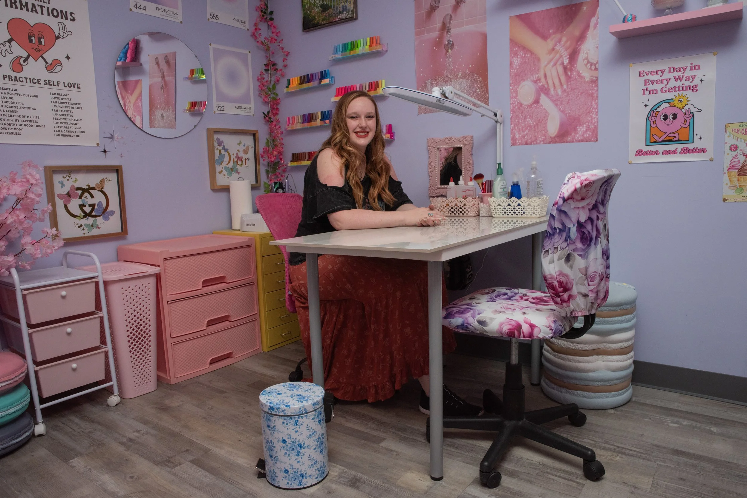 A woman with long wavy hair sitting at a nail salon station, smiling. The room is decorated with pink and purple accents, shelves with nail polish, framed artwork, posters with positive messages, and storage units. There is a patterned chair and a small round blue floral stool in the foreground.