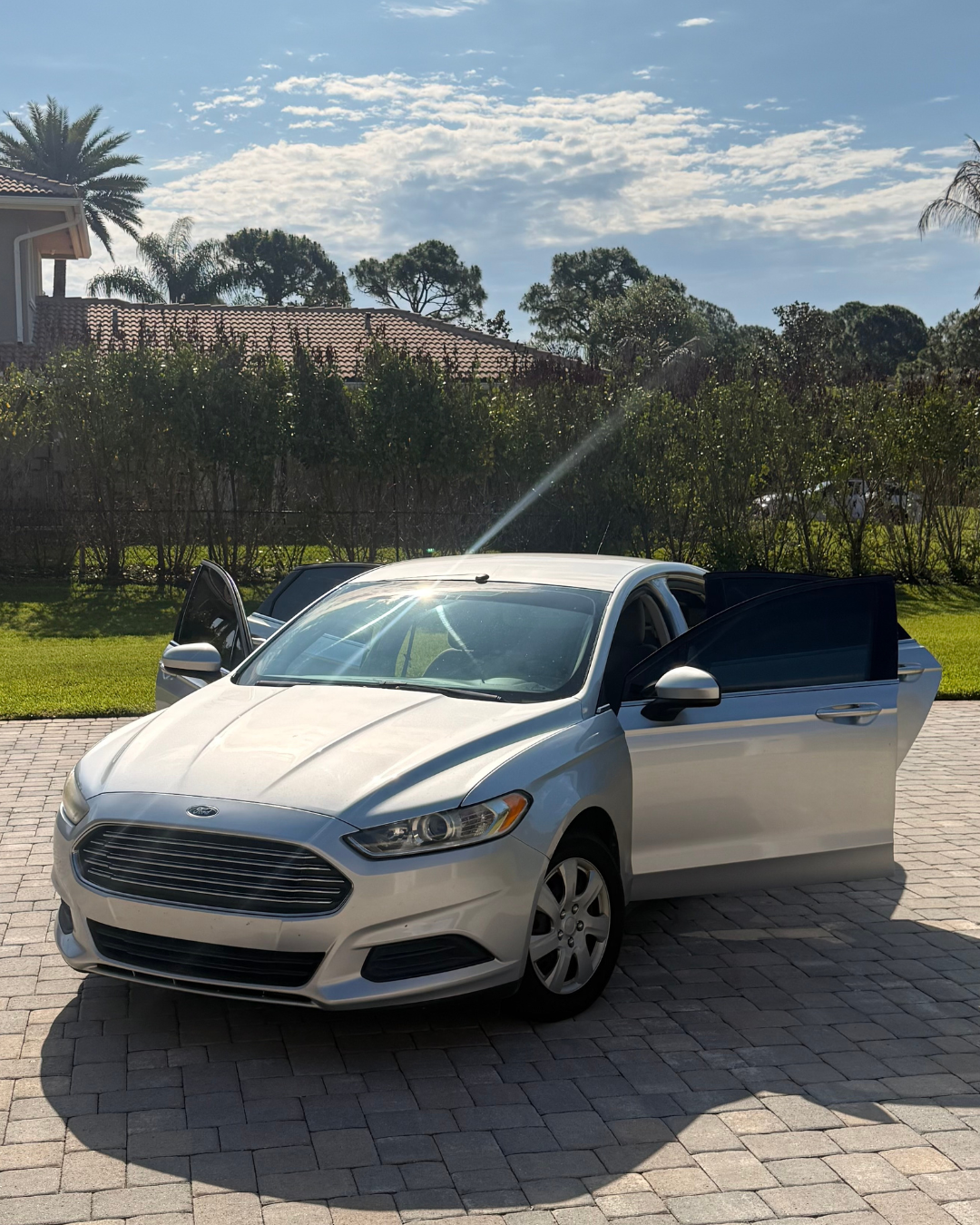 A silver sedan with its front and rear doors open parked on a brick driveway under a partly cloudy sky, with a green lawn, bushes, trees, and a house with a red-tiled roof in the background.
