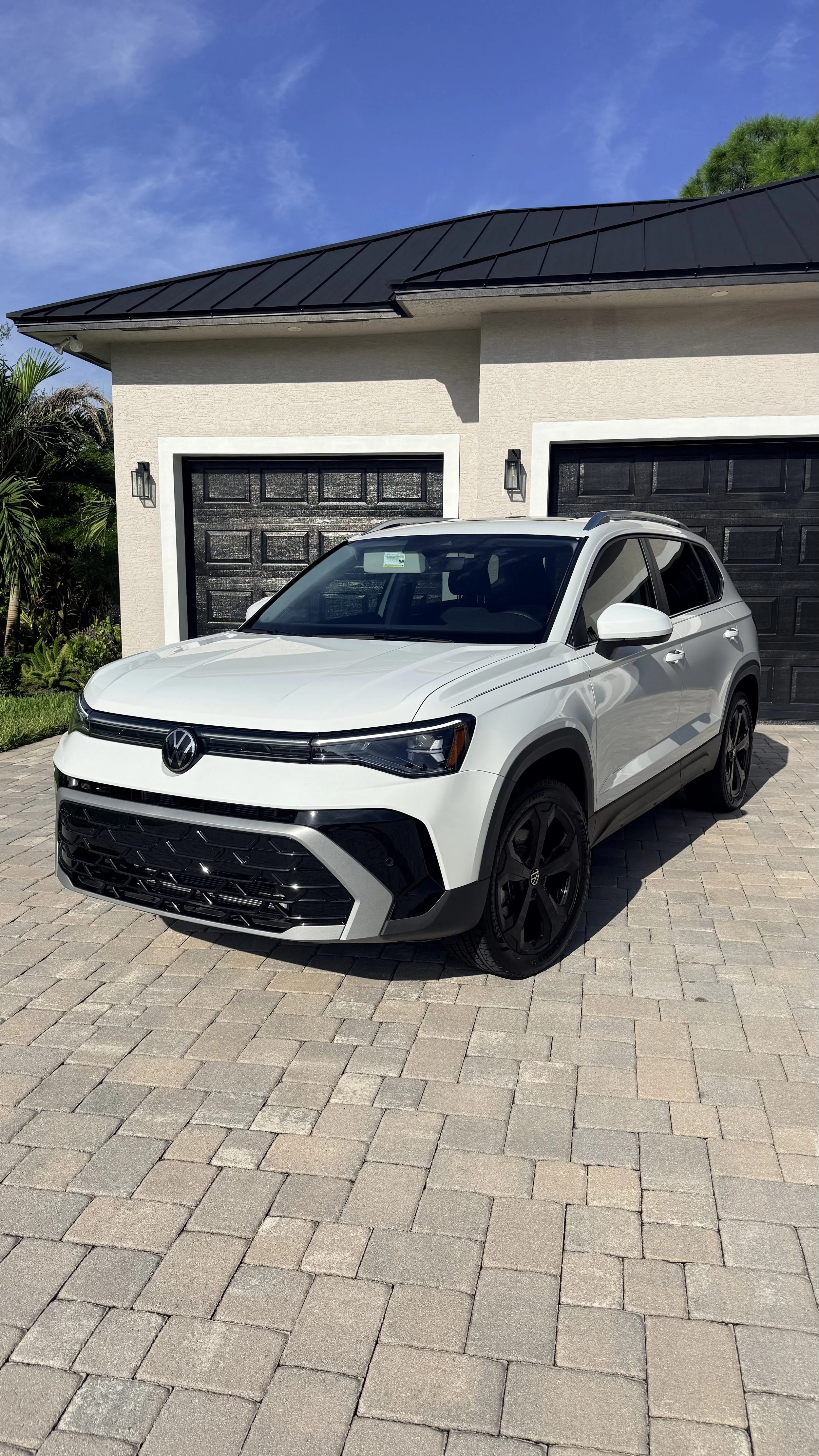 A white Volkswagen SUV parked in front of a house with black garage doors and a dark metal roof, with a paved driveway and some greenery on the side.