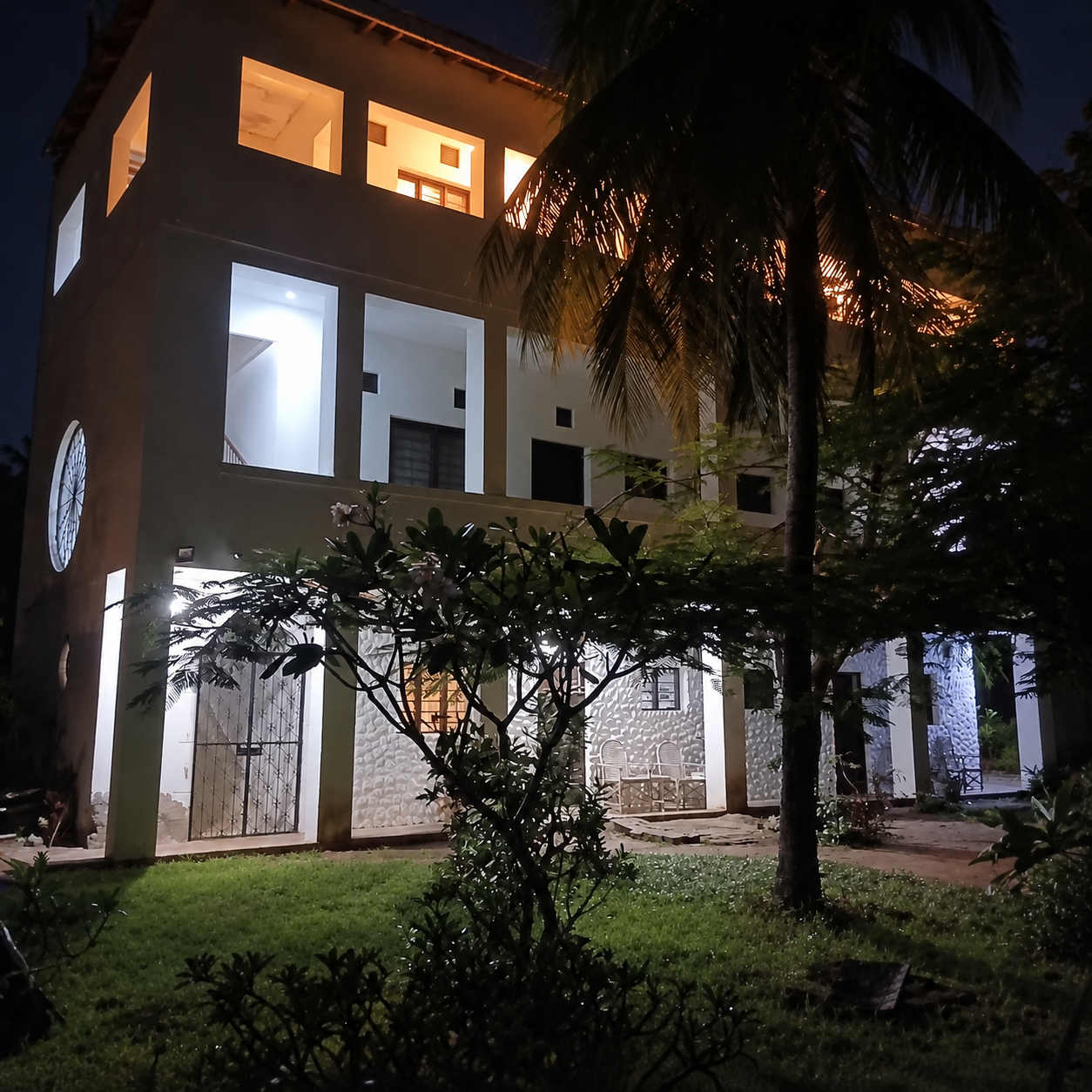 A modern multi-story house illuminated at night, with white walls, large windows, and trees in the foreground.