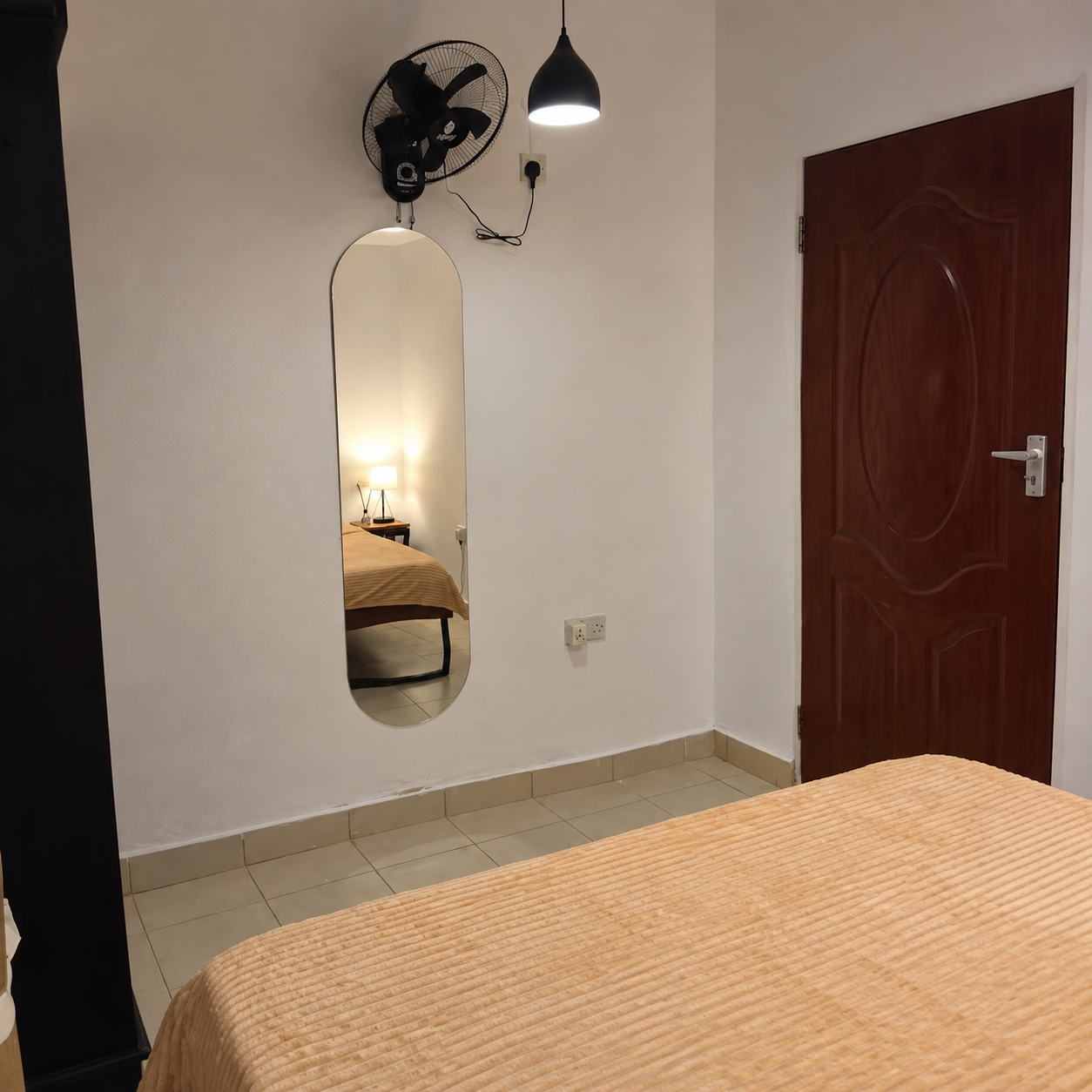 A bedroom with a bed covered in an orange bedspread, a mirror on the wall showing a reflection of a bedside lamp and bed, a wooden door, a wall-mounted fan, and a ceiling light.