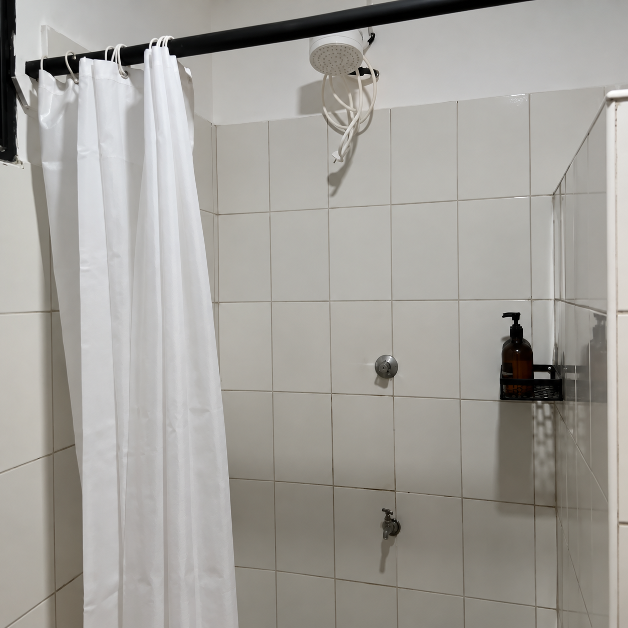 A simple bathroom shower area with white tiled walls, a white shower curtain, a ceramic showerhead, a small shelf with soap dispenser, and a faucet.