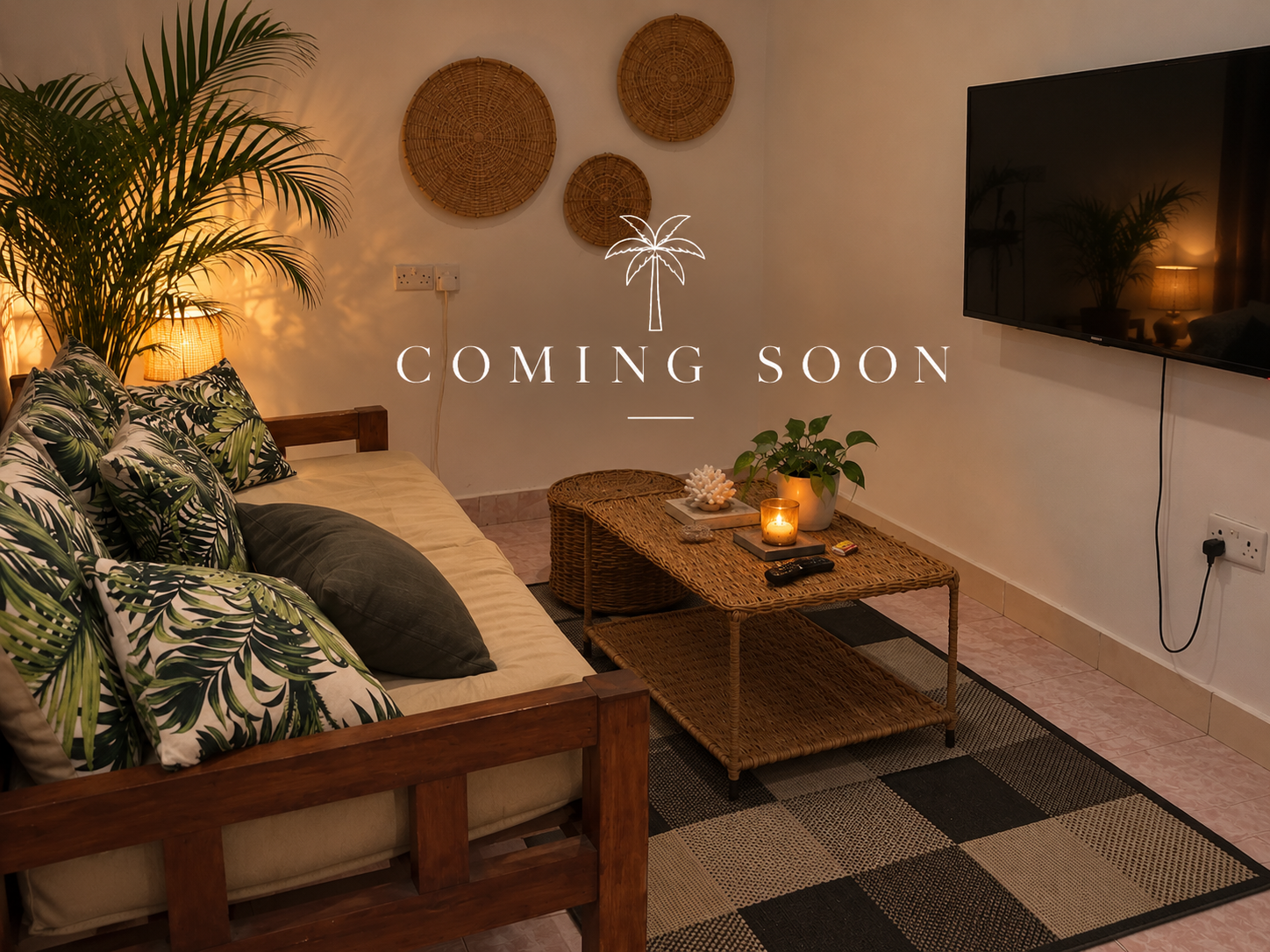 Living room with a wooden sofa that has tropical leaf patterned pillows, a wicker coffee table with a potted plant, candles, and decor. A TV is on the wall and a large plant in the corner. The photo has a 'Coming Soon' overlay.