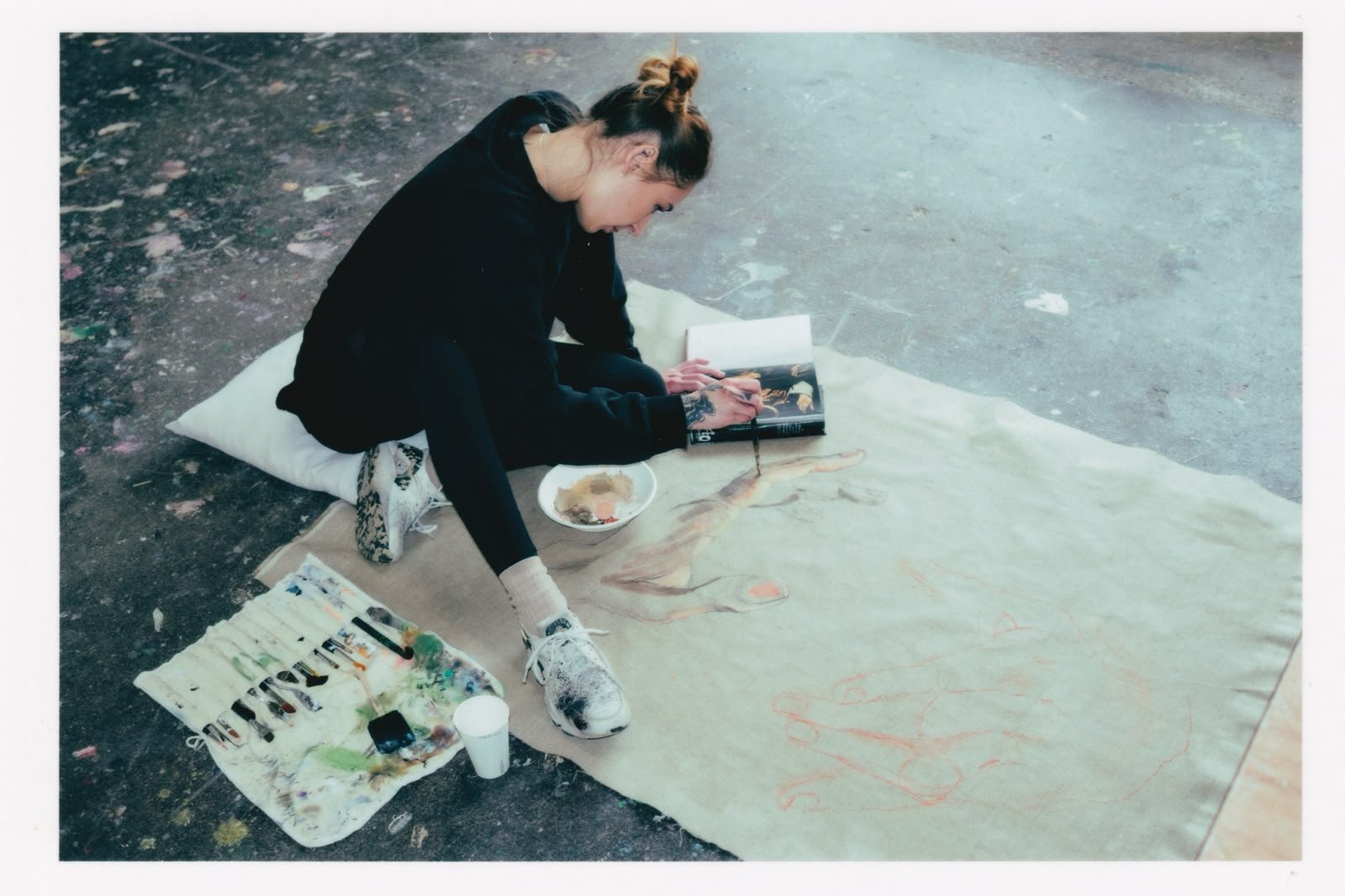 A woman painting on a large canvas outdoors while sitting on a cushion, surrounded by art supplies and a bowl of paint, with her hair tied in a bun and wearing casual clothes.