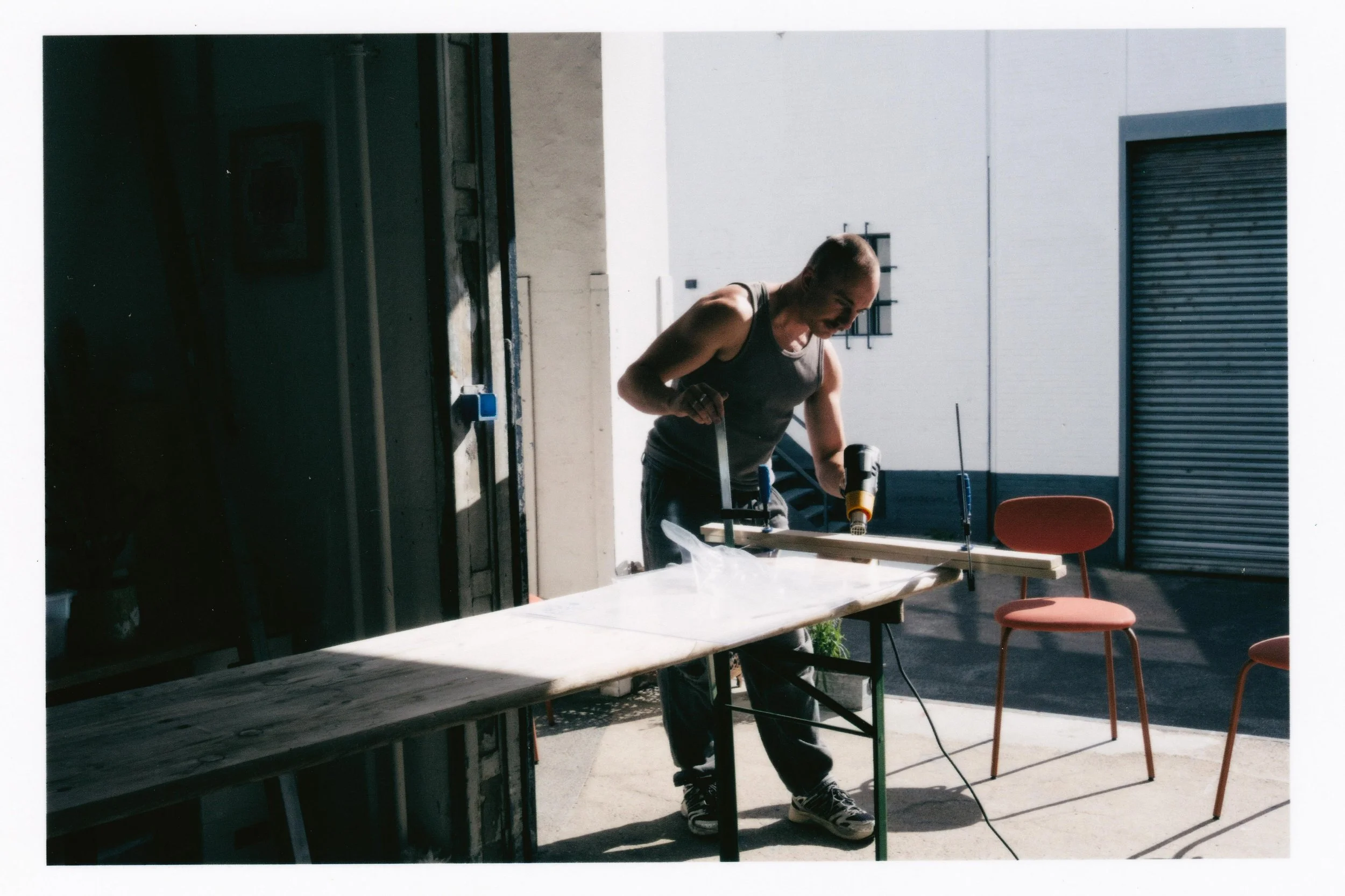 A man working with power tools at a table outside in a bright area, with chairs nearby and a large garage door in the background.