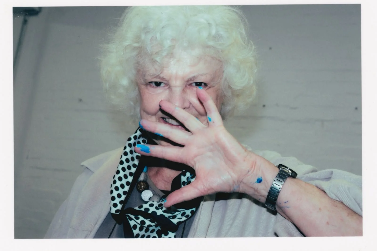 An elderly woman with curly white hair and light skin smiling at the camera, partially covering her face with her right hand which has blue paint on the fingernails, wearing a black and white polka dot scarf and a watch on her left wrist.