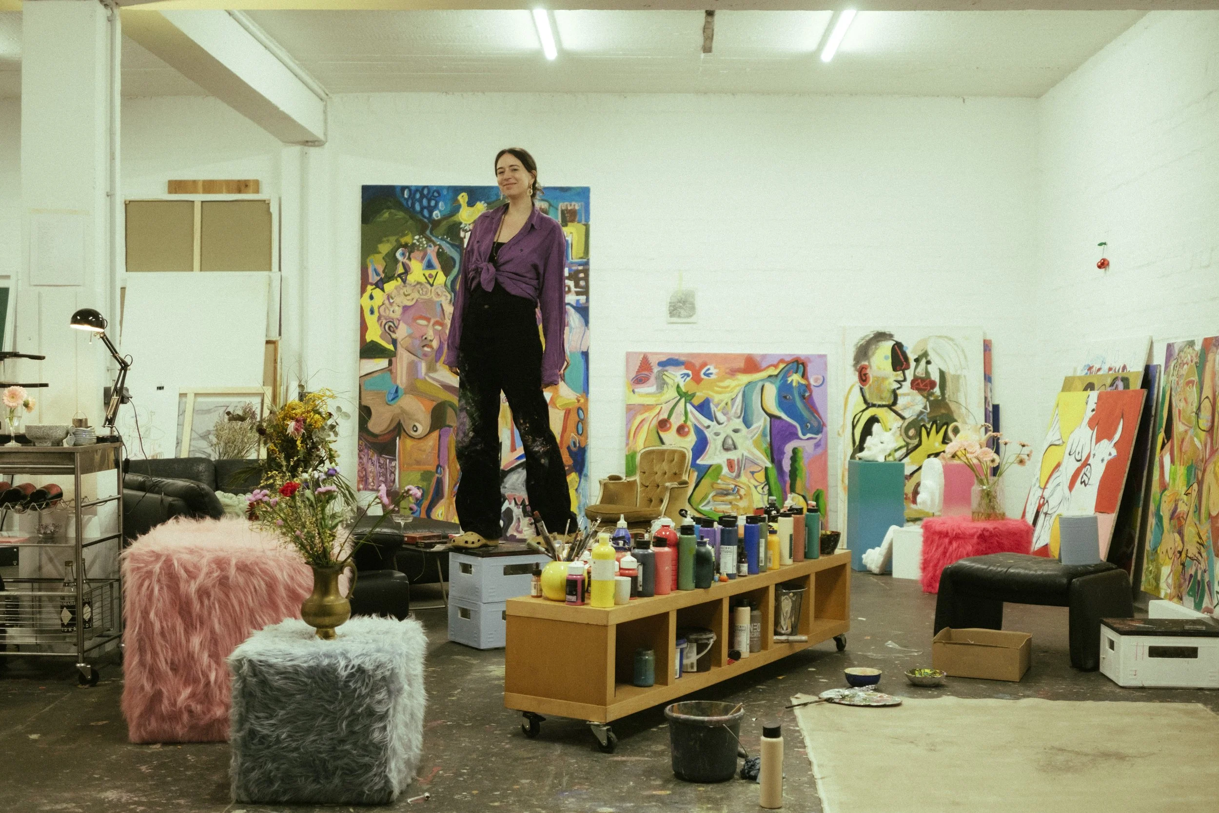 An artist standing in their colorful art studio surrounded by completed paintings, art supplies, and decorative furniture.