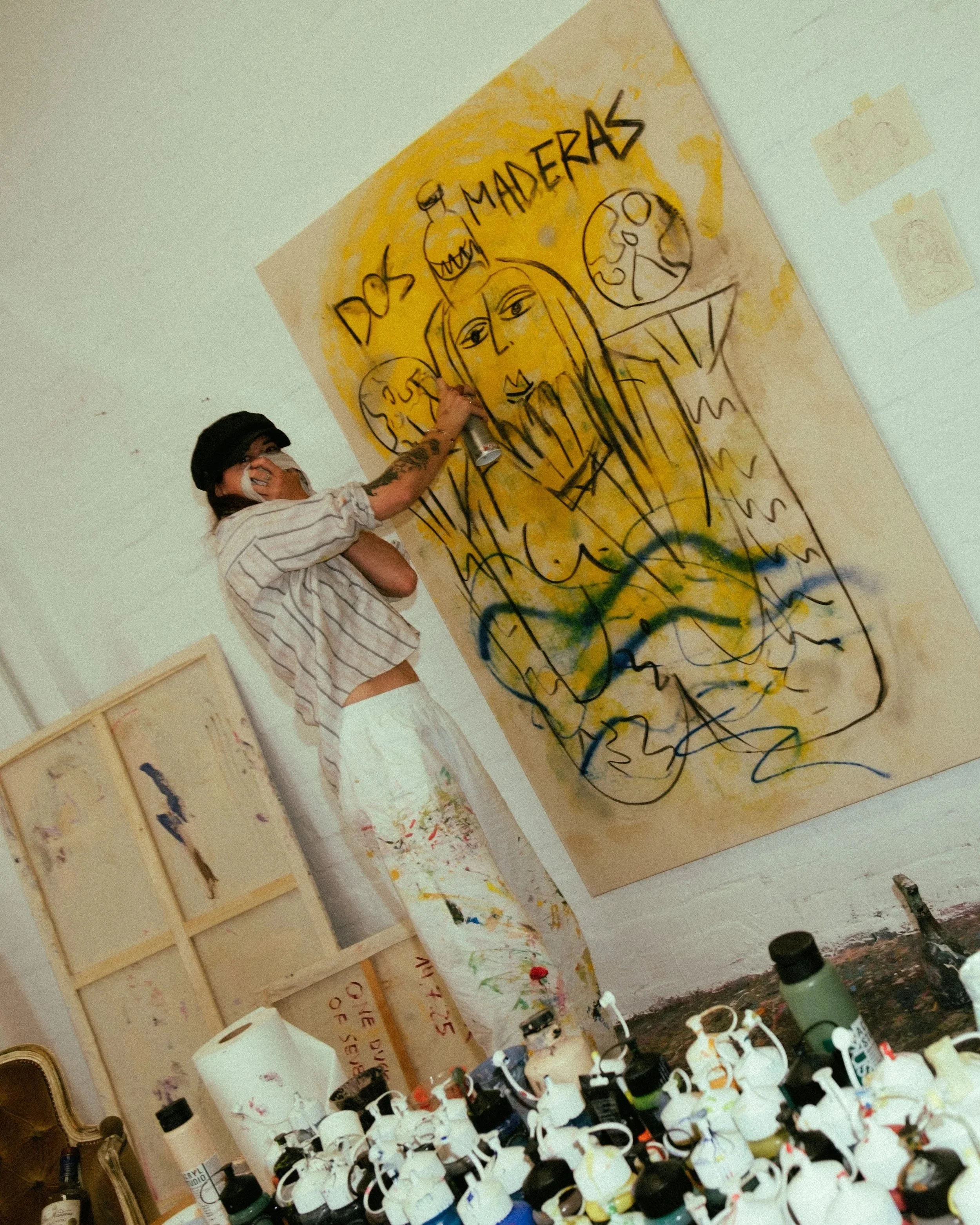 An artist creates abstract and colorful artworks on paper in a studio. The artworks include the words 'DOS' and 'MADEIRAS' with illustrations of faces and abstract shapes, with various paint bottles and supplies in the foreground.