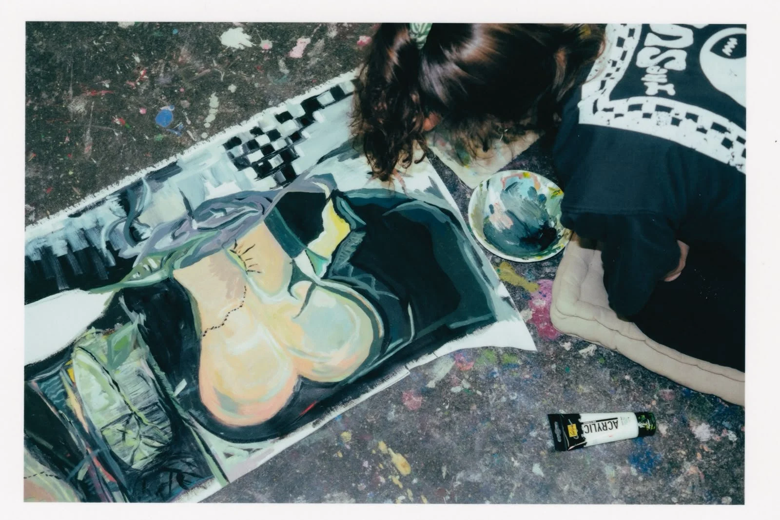An artist working on a colorful painting of a woman wearing sunglasses, with painting supplies and a palette nearby, sitting on a padded cushion on a speckled floor.
