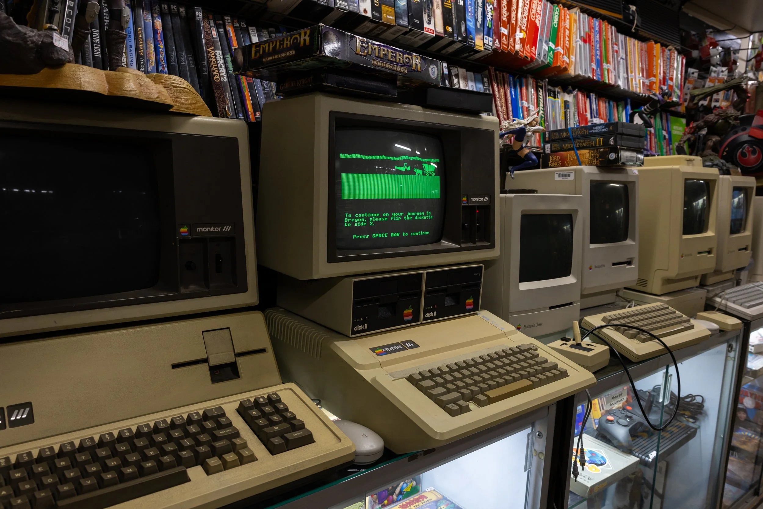 Several vintage Apple Macintosh computer monitors, keyboards, and accessories are displayed on glass shelves at a store. One monitor shows a green monochrome screen with a loading message. The background features shelves filled with board games and other collectibles.