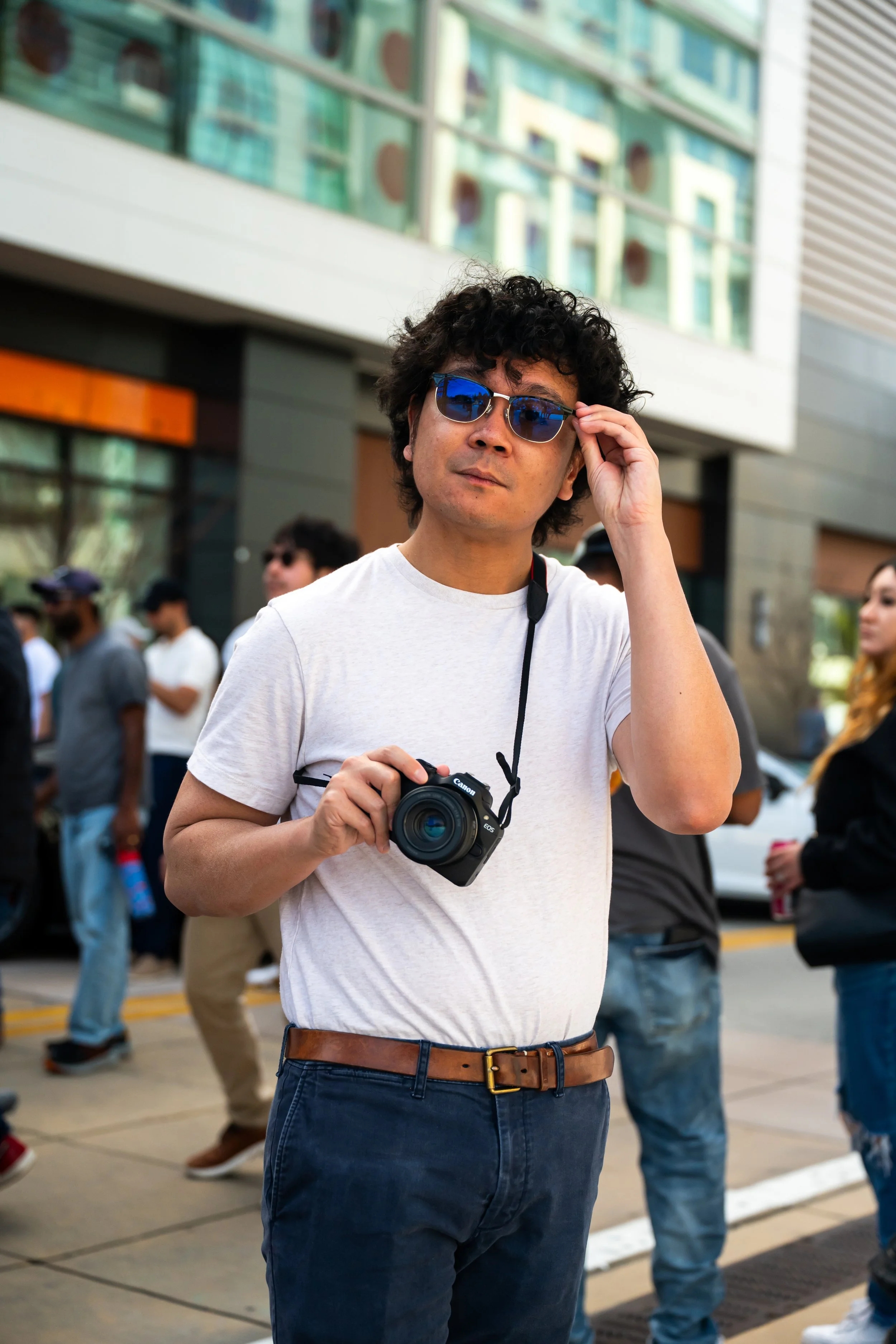 A man with curly black hair and sunglasses is holding a camera in his left hand while adjusting his glasses with his right hand in a busy outdoor area with people and modern buildings in the background.