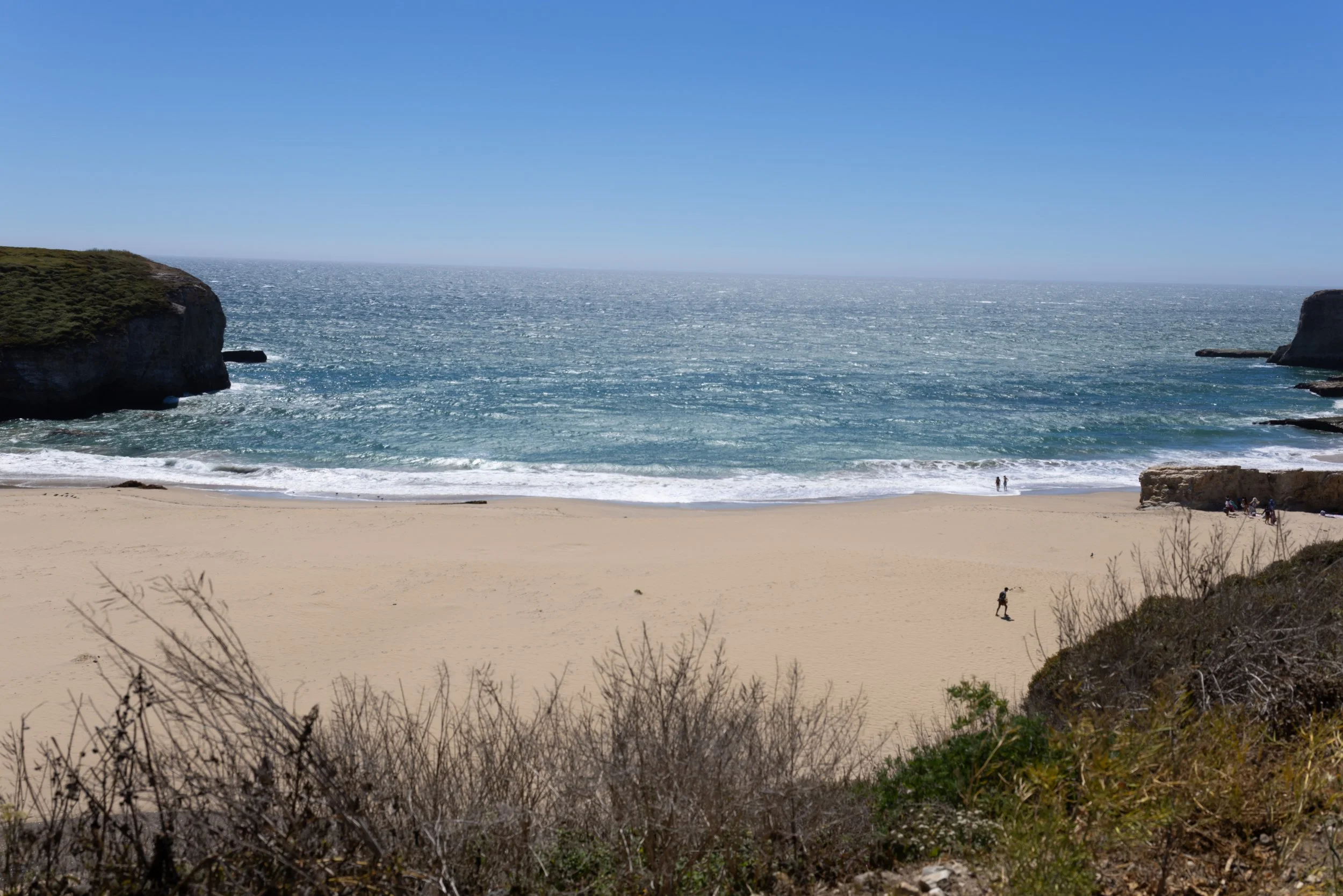 A sandy beach with a few people walking near the shoreline, surrounded by grassy dunes and cliffs on the left and right, with the ocean and a clear blue sky in the background.