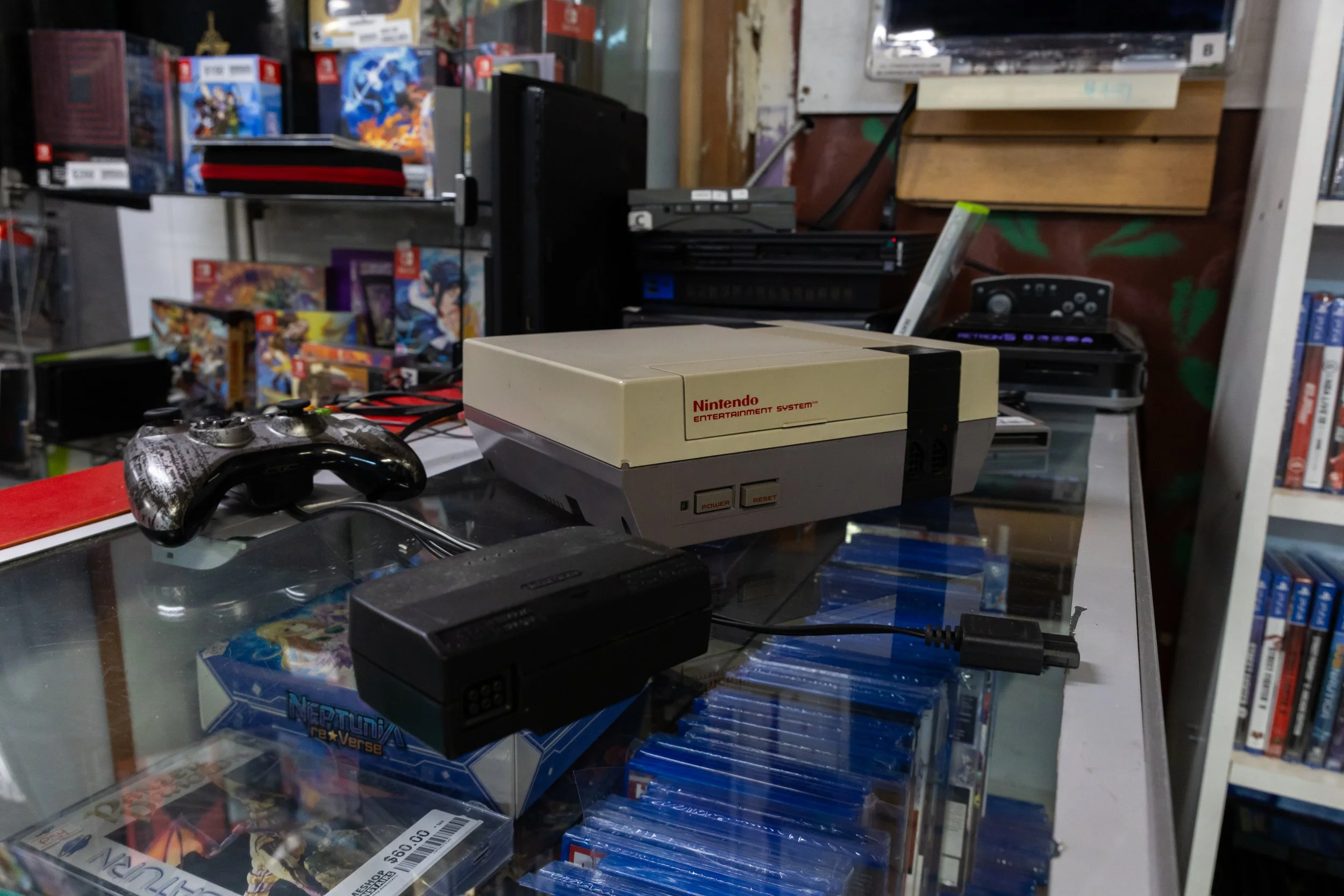 A collection of video games and gaming accessories displayed on shelves and a glass table. Prominent items include a vintage Nintendo Entertainment System, various game boxes, controllers, and gaming-related items.