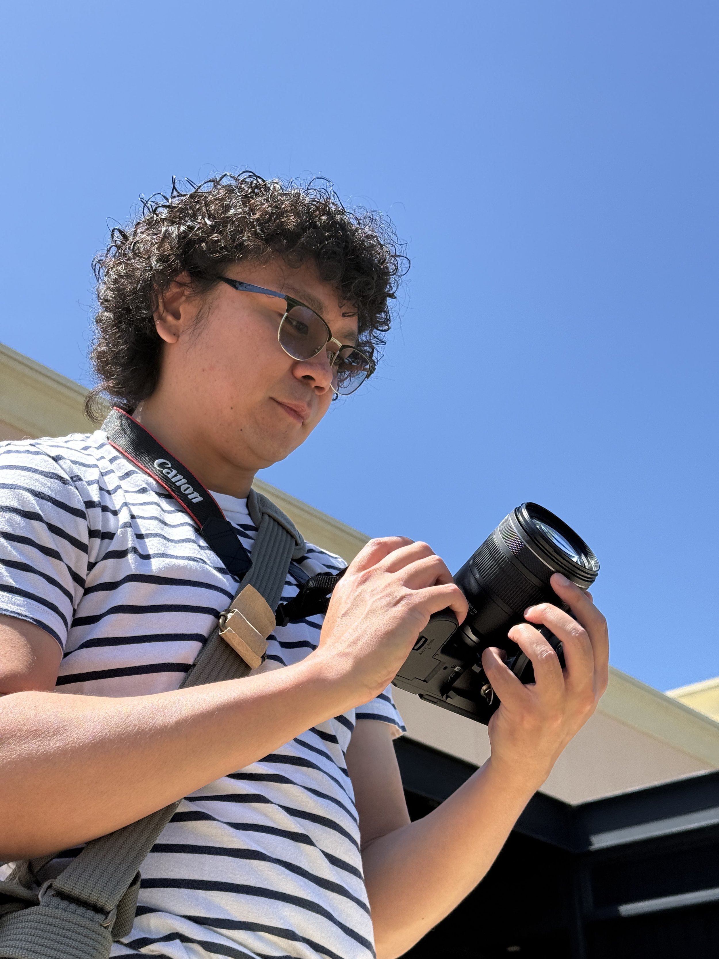 A young man with curly hair, glasses, and a striped shirt holding a digital camera, looking down at it, outdoors against a bright blue sky.