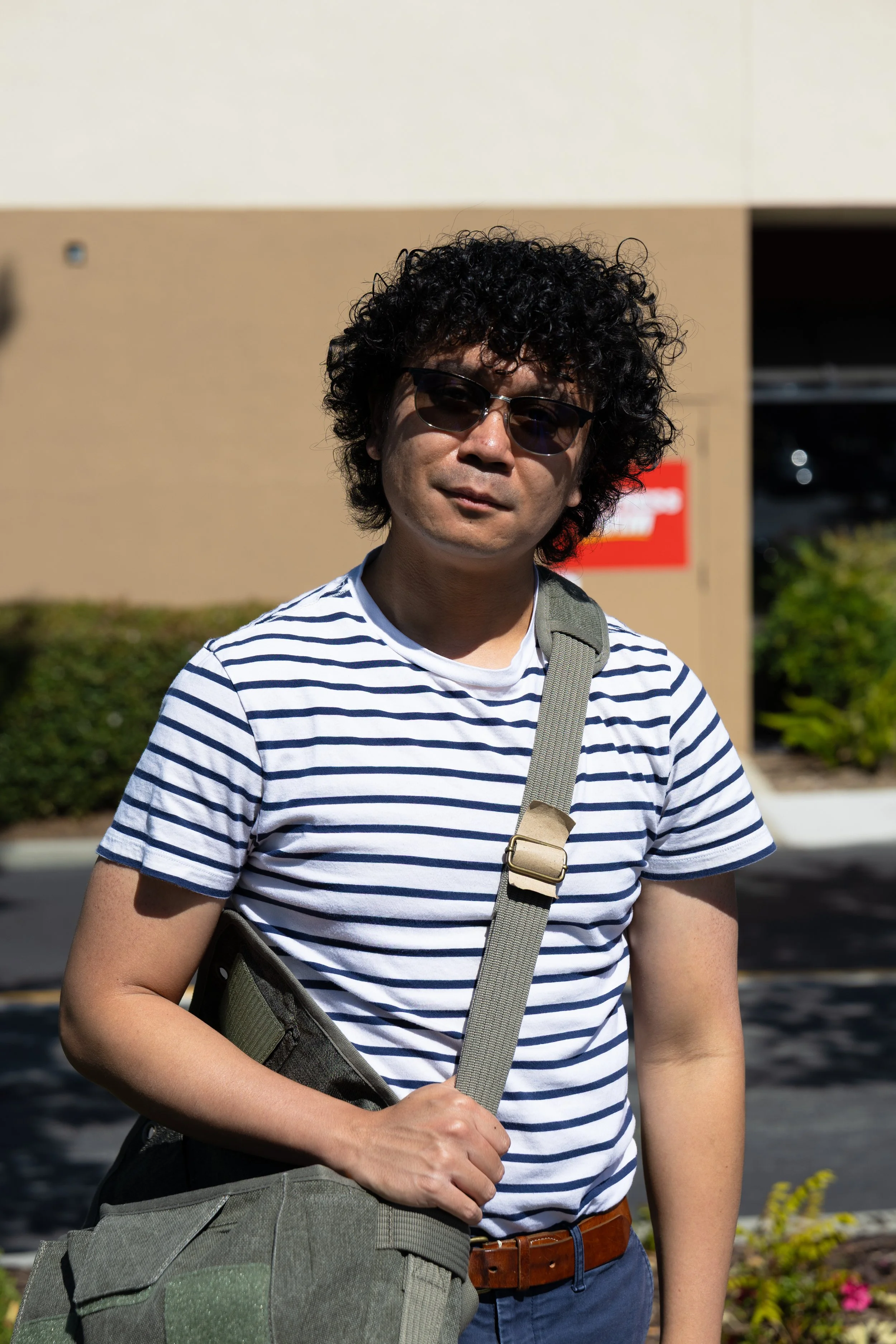 A man with curly black hair, wearing dark sunglasses, a white and navy striped t-shirt, and carrying a gray shoulder bag, standing outside in bright sunlight with a building and a red sign in the background.