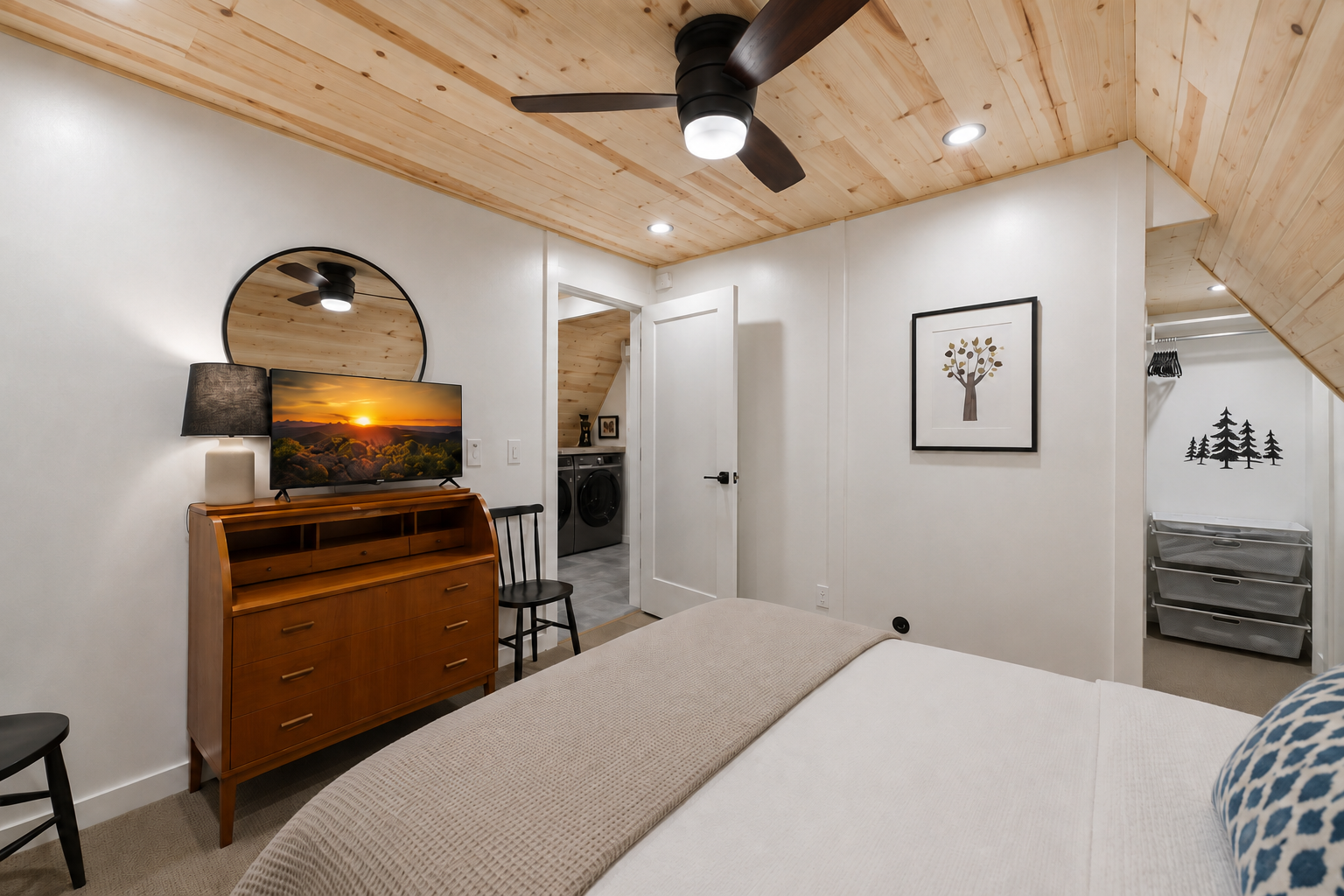 A cozy bedroom with a wooden ceiling, white walls, and minimalist decor. There is a bed with a beige blanket, a black chair, a wooden dresser with a TV and a lamp, and wall art depicting trees. An open door shows a laundry area with a washing machine