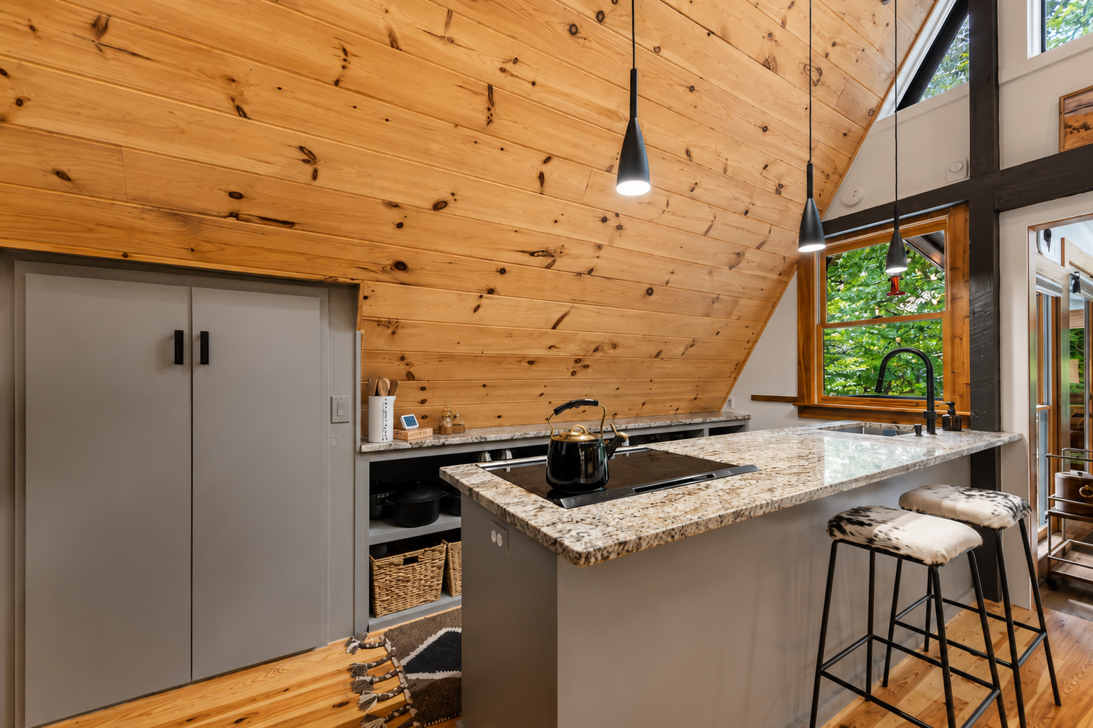 Kitchen with wooden sloped ceiling, granite countertop island, black kettle, black sink and faucet, backsplash with open shelves, two barstools with cowhide seats, large window showing green foliage outside, black pendant lights hanging above island,