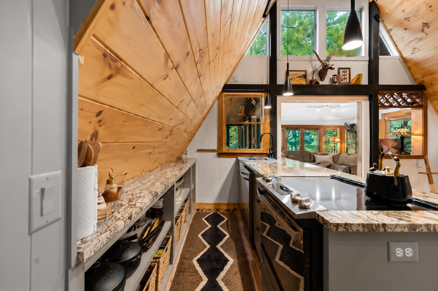 View of a modern kitchen with a granite countertop, black stove, and a black kettle. Wood-paneled ceiling and walls, open shelves with baskets and kitchenware, and a living room with a beige sectional sofa and a lamp visible through an open doorway.