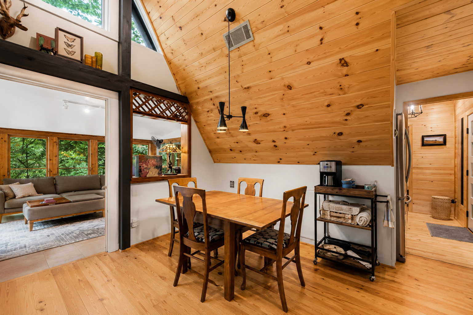 A dining area with a wooden table and four chairs, situated under a vaulted wooden ceiling with a hanging light fixture. To the right, there is a rolling cart with a coffee maker and supplies, next to a refrigerator. An open doorway leads to a living