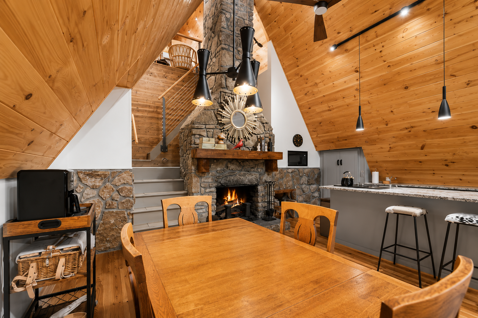 Interior view of a cozy room with wood-paneled sloped ceiling, stone fireplace with fire, wooden dining table with chairs, and kitchen area with bar stools, black pendant lights, and countertop.