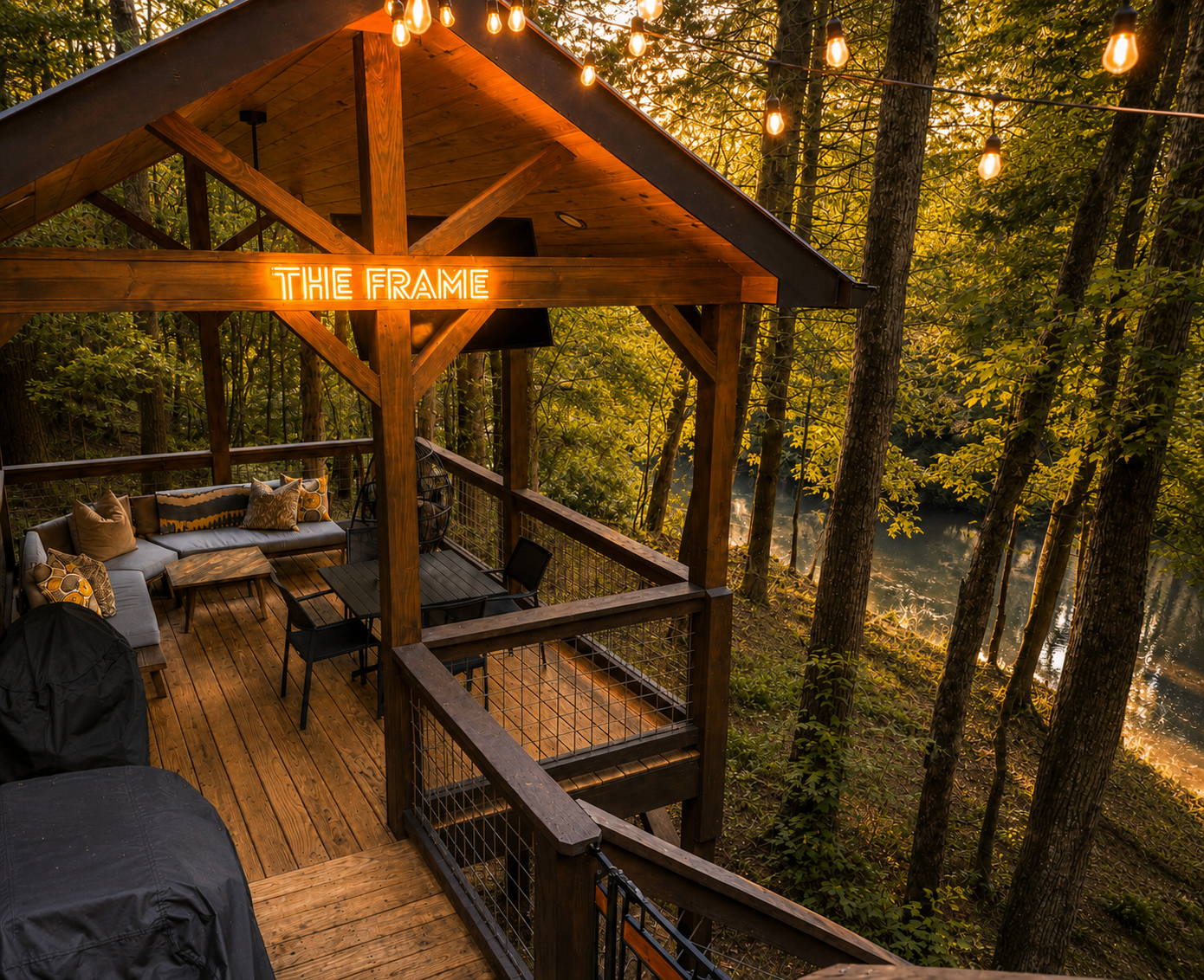 A wooden outdoor deck with patio furniture, cushions, and a neon sign that reads 'THE FRAME', surrounded by trees and a body of water in the background, illuminated by string lights at sunset.