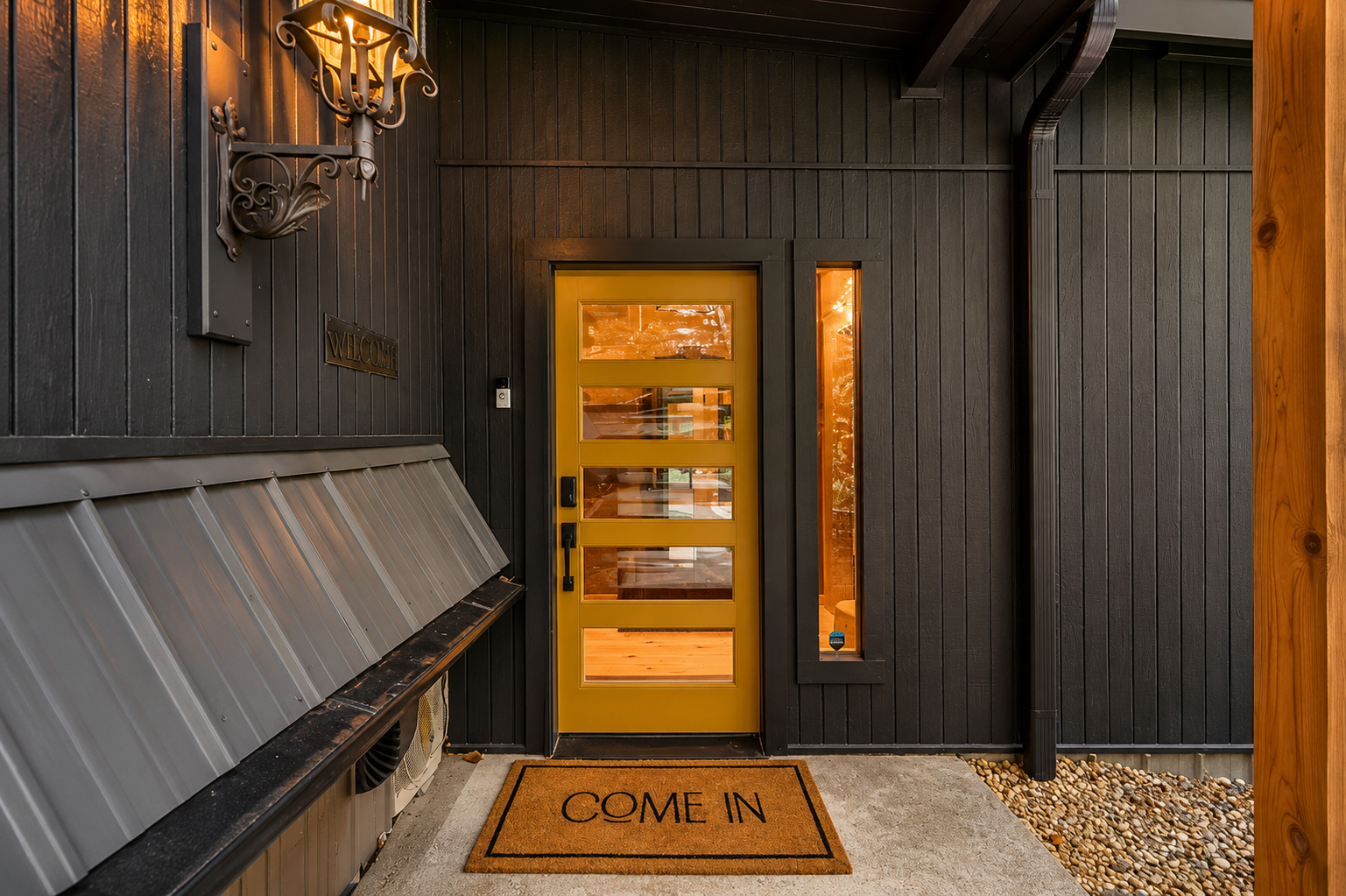 Front porch area of a house with a yellow door, black wooden siding, and a welcome mat that says 'COME IN'. The porch has a wall sconce, a narrow side window, and a metal awning above a small utility area.