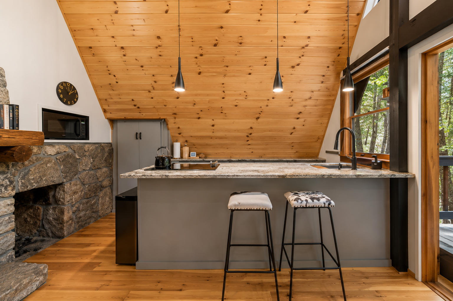 Kitchen with stone and wood accents, a marble countertop island with two stools, black pendant lights, a window showing trees outside, and appliances including a microwave and coffee maker.