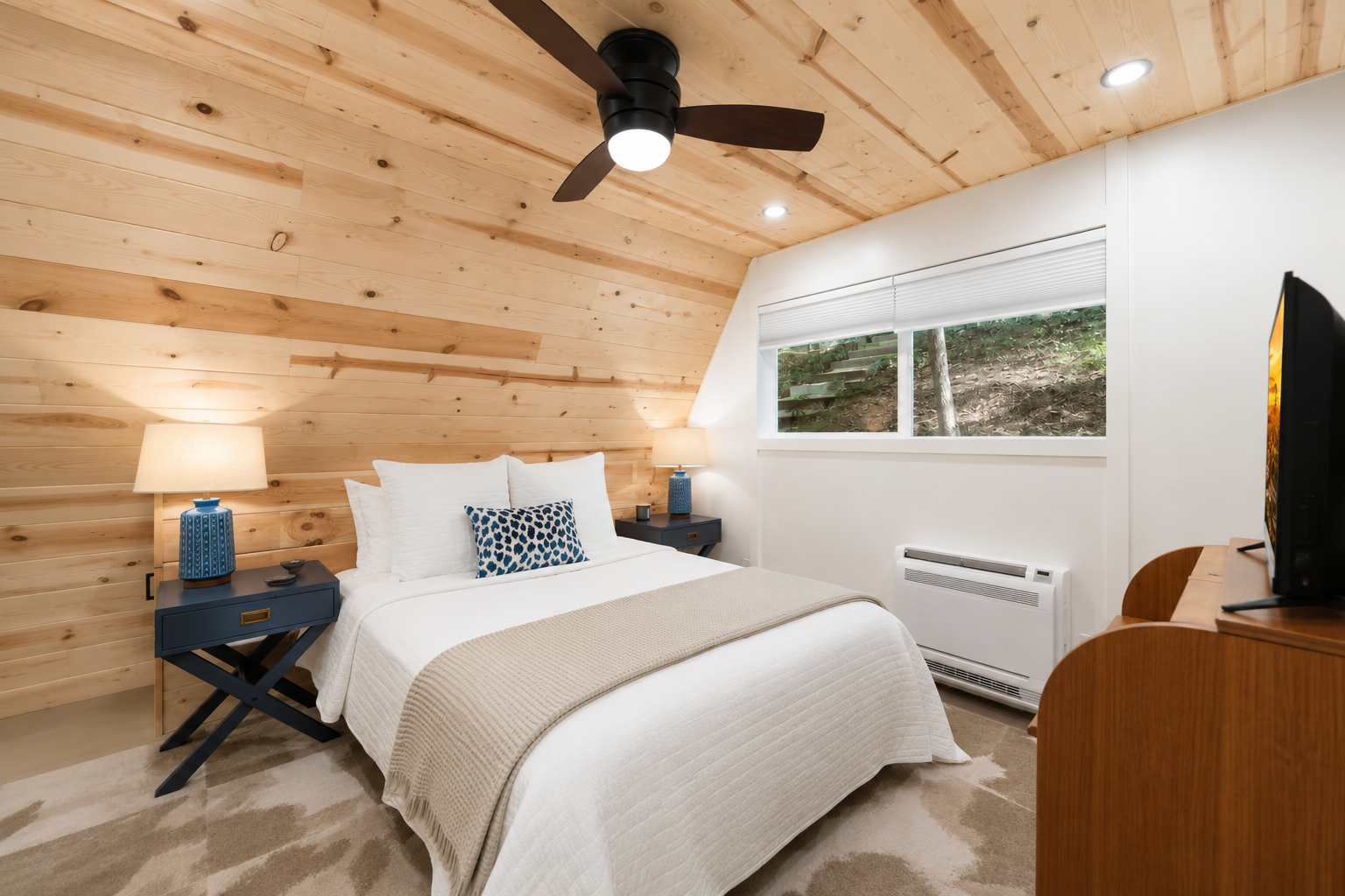 A cozy bedroom with a wooden sloped ceiling, white walls, a white bed with multiple pillows, two blue nightstands with lamps, a window revealing a wooded outdoor area, a TV on a wooden dresser, and a ceiling fan.