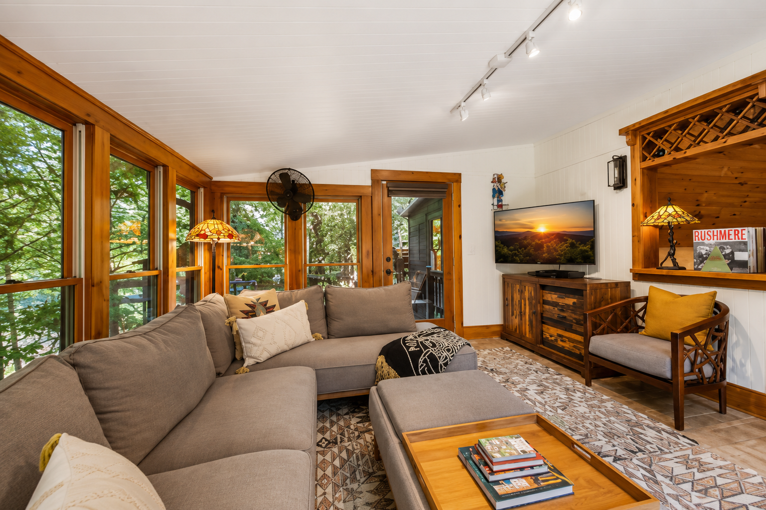 Living room with large windows, gray sectional sofa with throw pillows, wooden accents, mounted flat-screen TV, armchair, coffee table with magazines, and decorative lamps