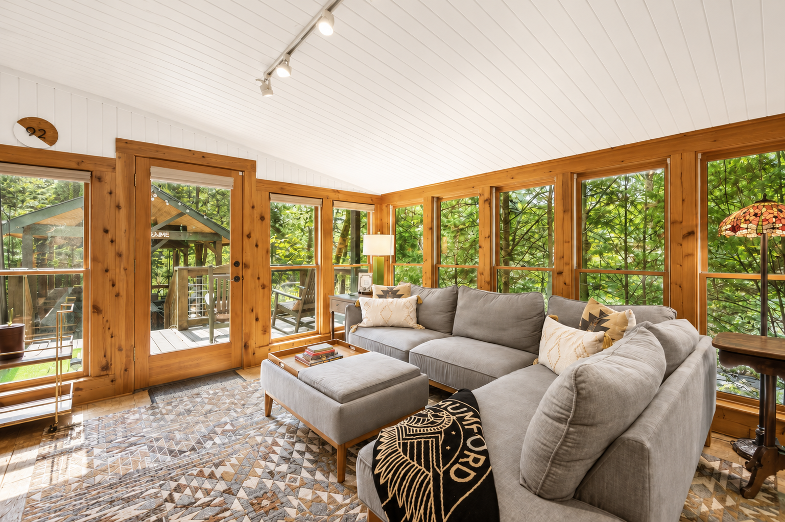 Living room with large windows showing green trees outside, a gray sectional sofa with decorative pillows, a wooden door leading to an outdoor deck, a patterned area rug, a side table with a lamp, and a stained glass lamp in the corner.